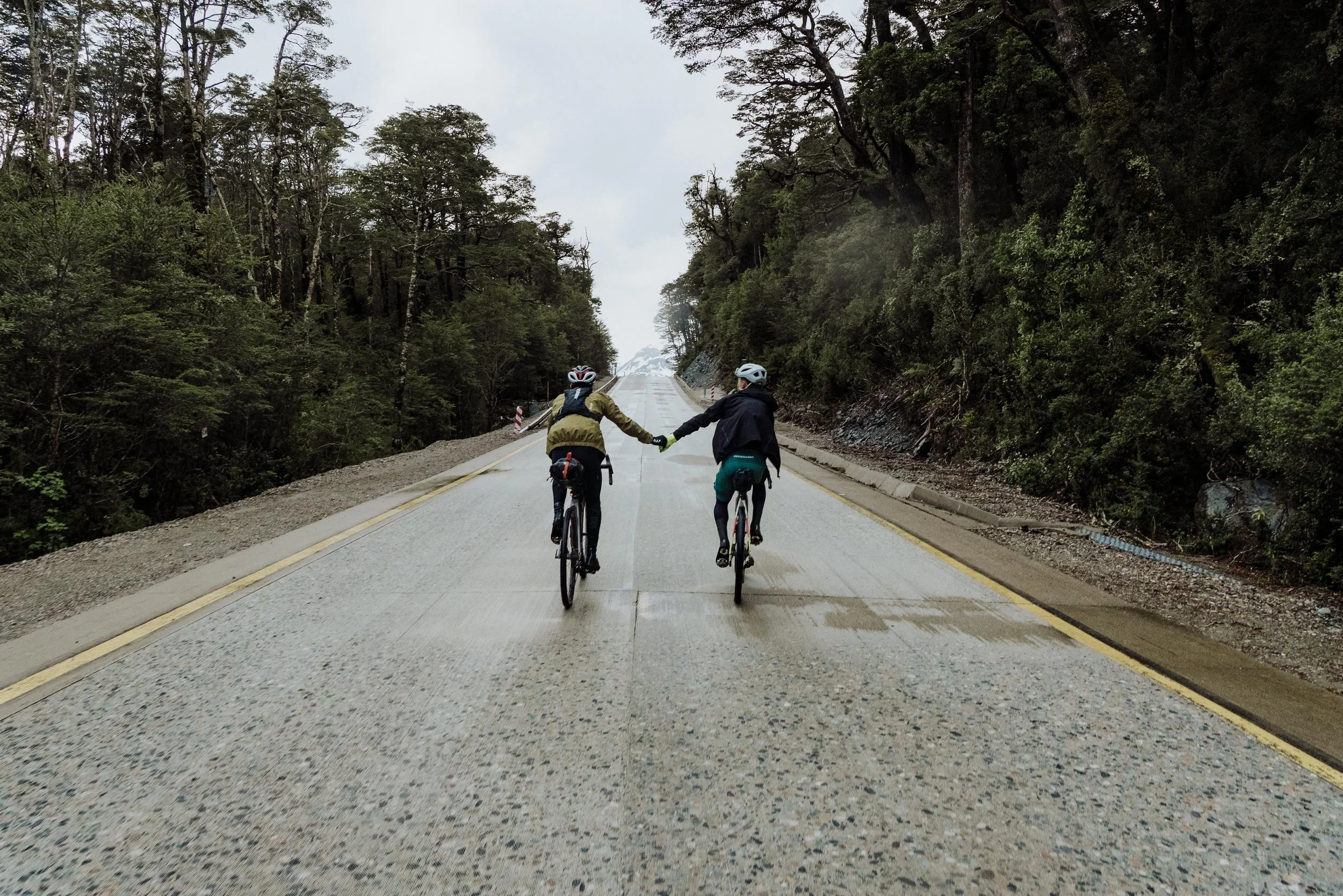 Two people riding bicycles on a wet, empty road surrounded by trees, holding hands, during overcast weather.