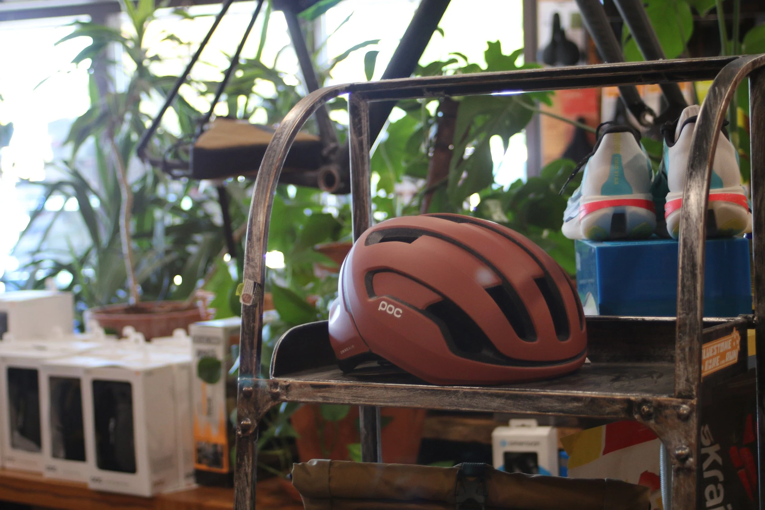 A brown bicycle helmet with black vents resting on a metal shelf, with a pair of sneakers hanging above on the same shelf. The background includes potted plants and various boxes.