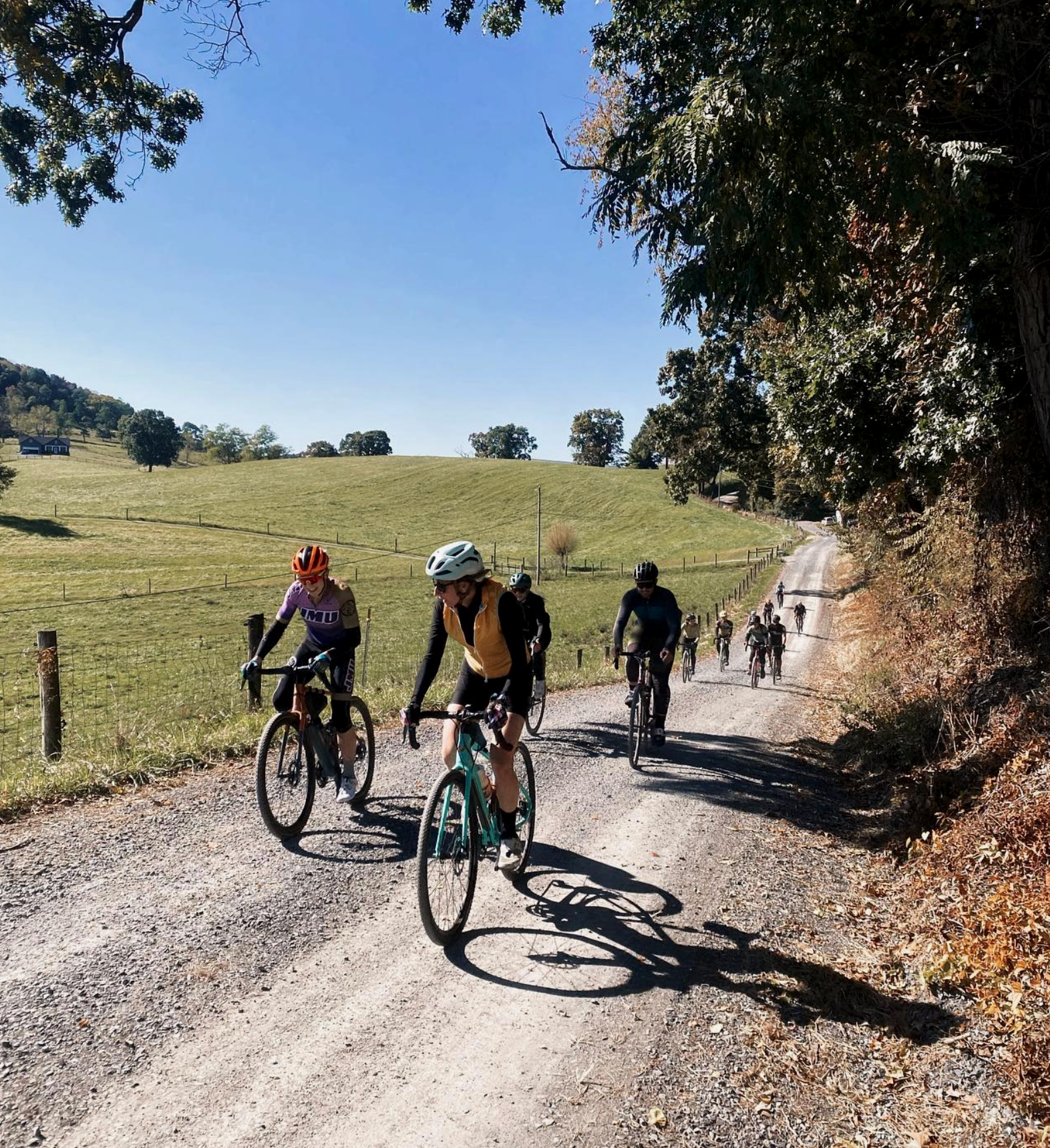 A group of cyclists riding on a gravel trail through a rural landscape with green fields, trees, and a clear blue sky.