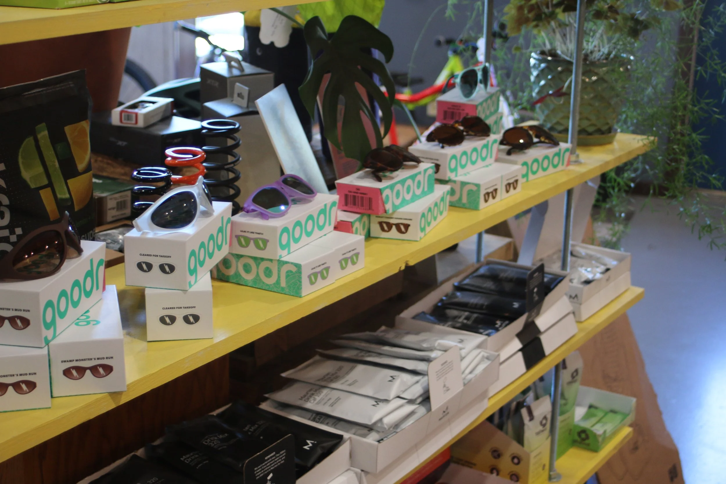 Display of sunglasses and accessories on yellow shelving in a retail store, with plants and merchandise in the background.