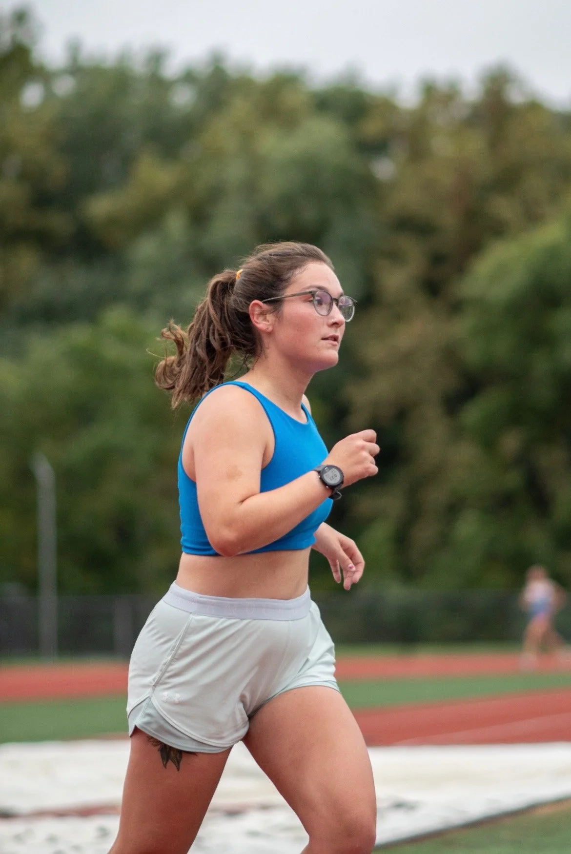 A woman running on a track wearing a blue sports bra, white shorts, glasses, and a watch, with a background of trees and a cloudy sky.