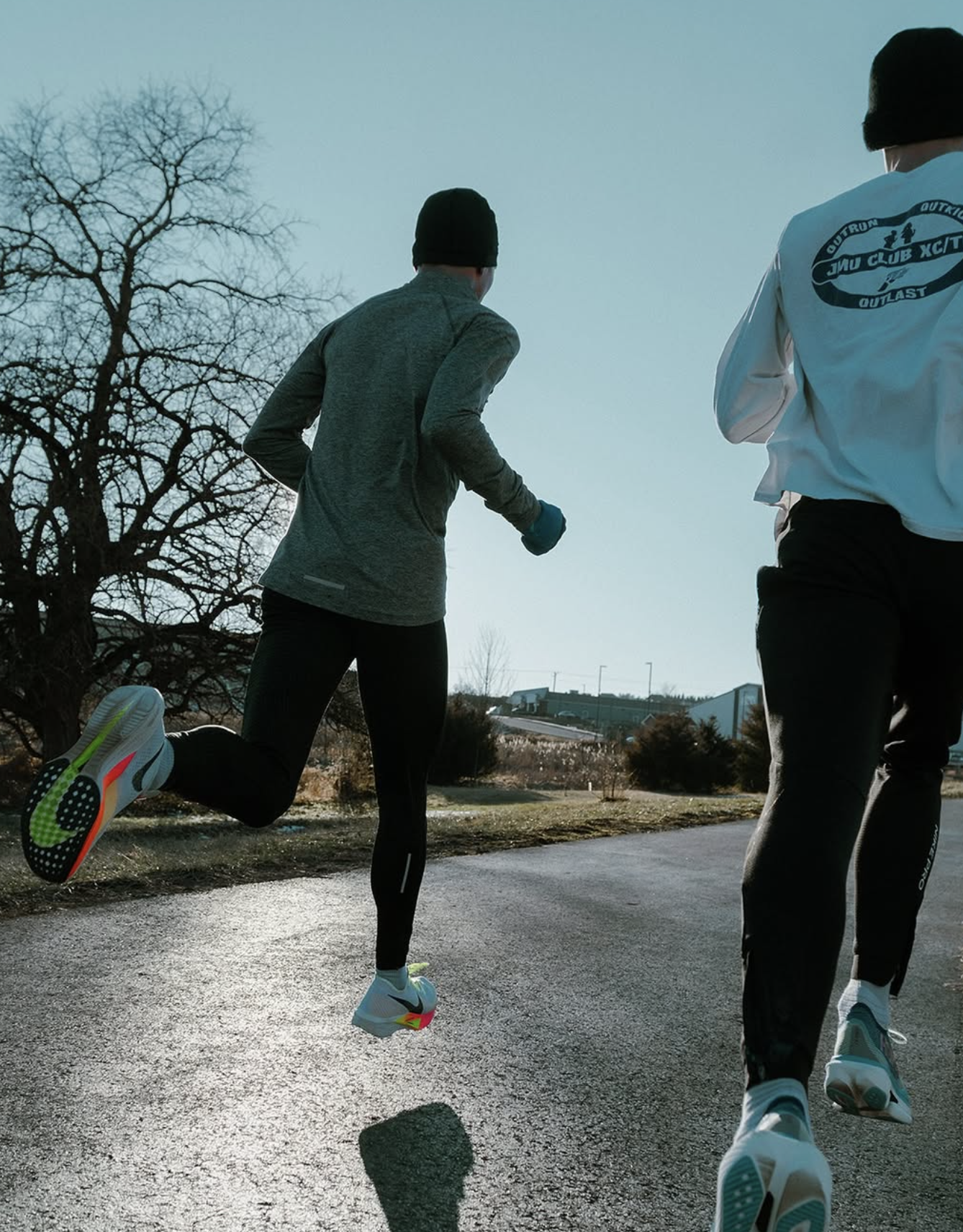 Two people running outdoors on a paved path during daytime, wearing athletic clothing and beanies, with trees and buildings in the background.