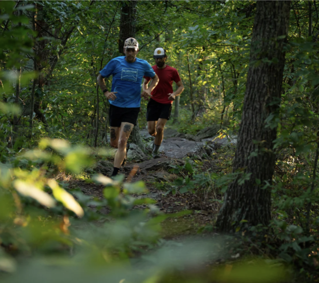 Two men trail running on a forest trail surrounded by green trees and foliage.