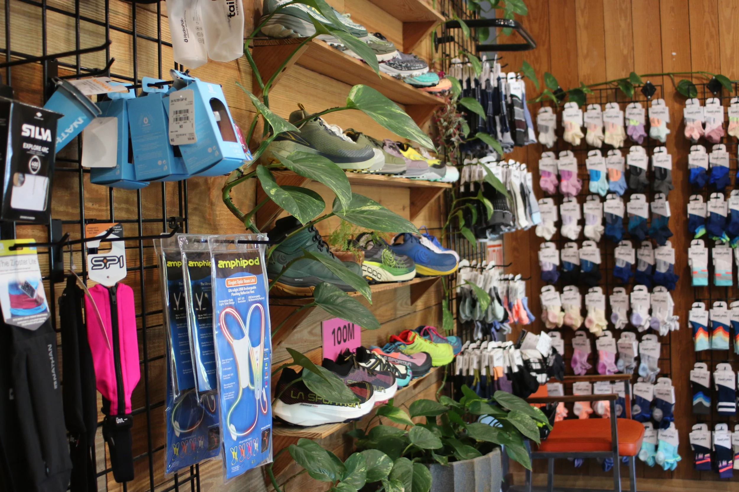 A retail store display with athletic shoes on wooden shelves, various socks and accessories hanging on hooks, and potted green plants on the wood-paneled wall.