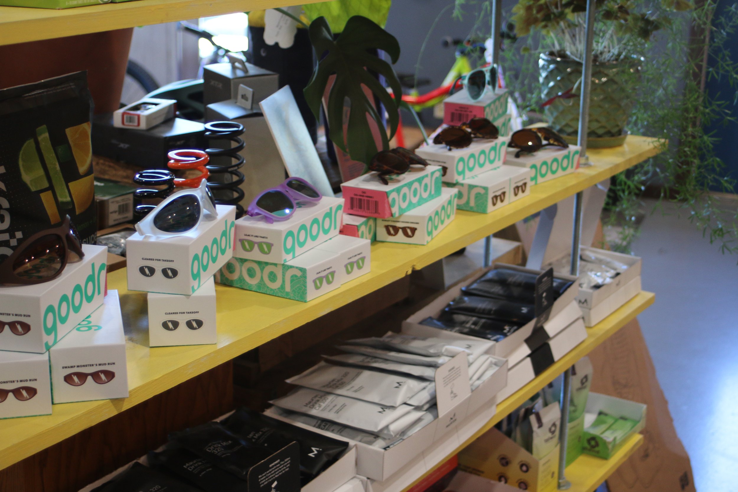 Display of sunglasses and accessories on a yellow retail shelf, with various boxes and plants in the background.