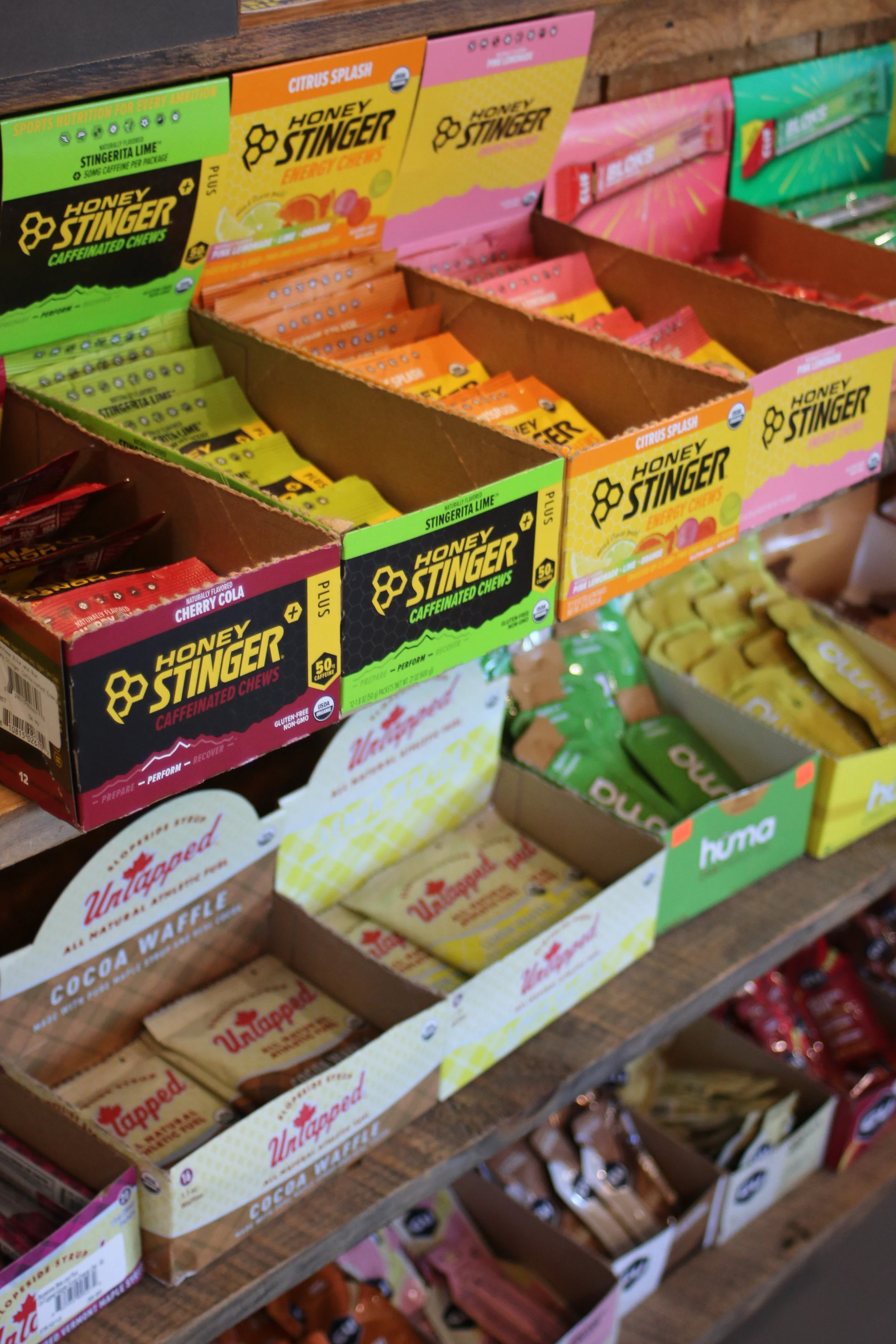 Display of Honey Stinger energy chews in various flavors, including Citrus Splash, Cherry Cola, and Stinger Lime, arranged in open boxes on a wooden shelf.