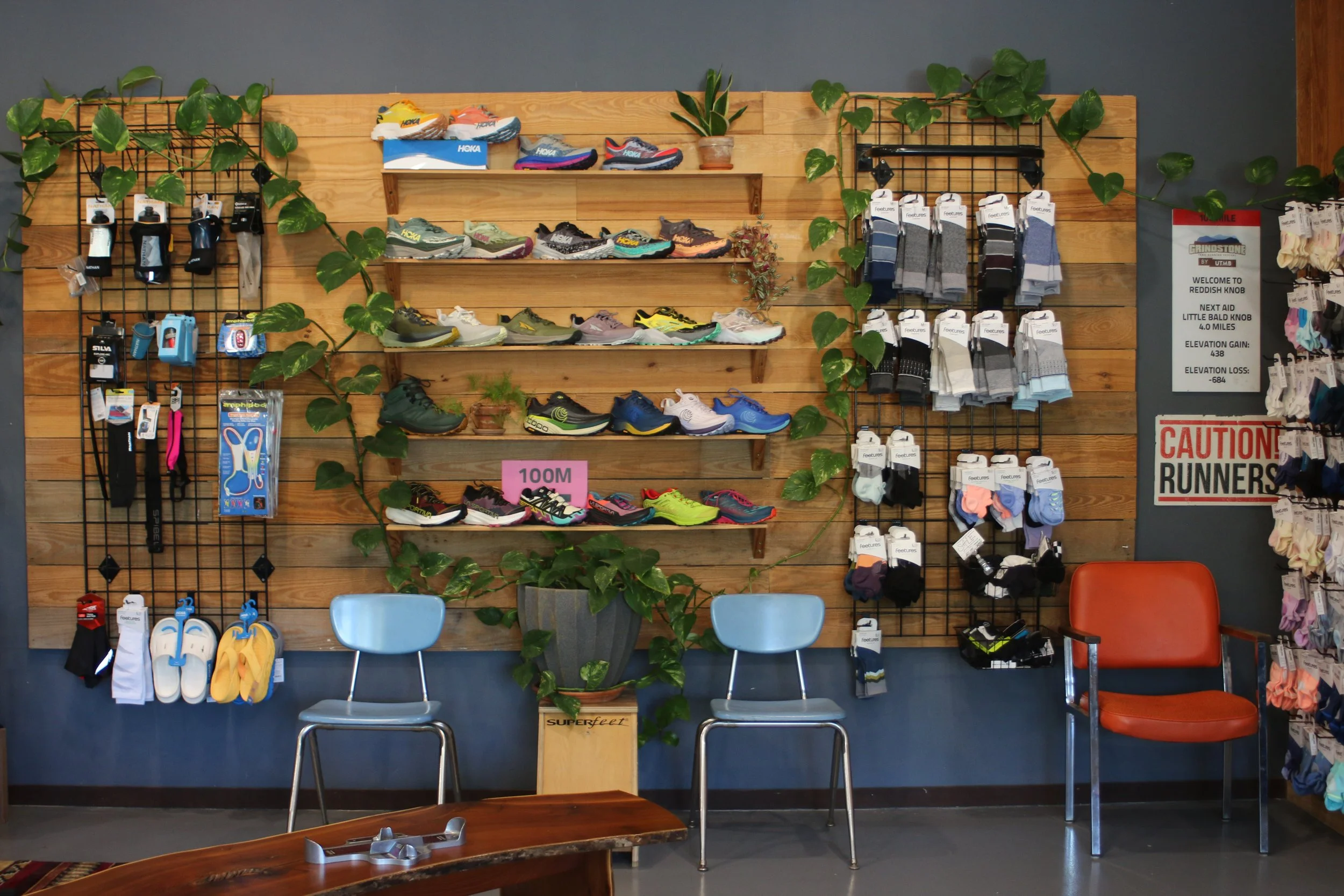 Display of running shoes, socks, and accessories on a wooden wall at a sporting goods store with chairs and plants in front.