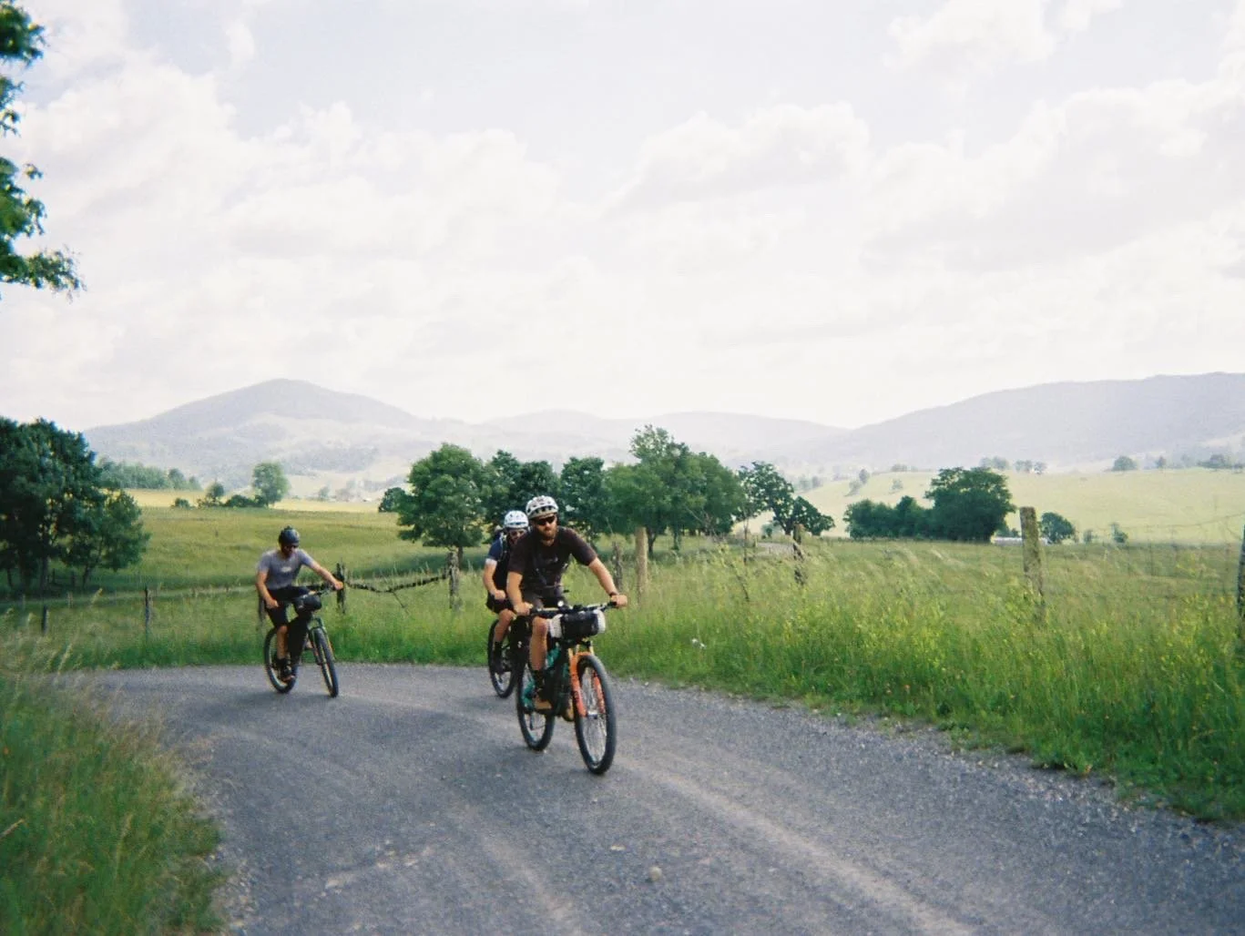 Three bikers in helmets riding on a gravel country road with green fields, trees, and mountains in the background under a partly cloudy sky.