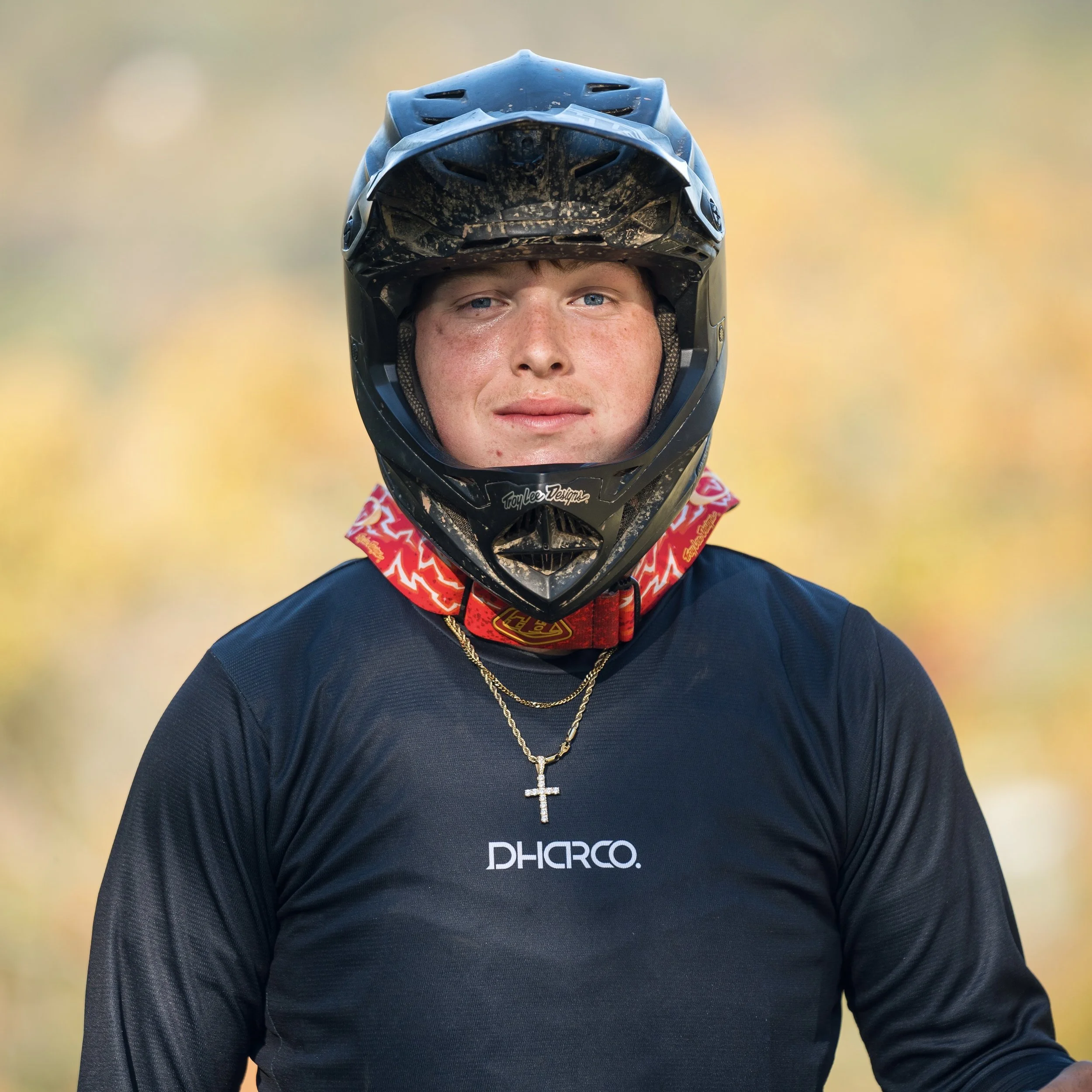 A young man wearing a black motocross helmet and a black long sleeve shirt with 'Dharco' logo, a red neck gaiter, a gold chain with a cross, and standing outdoors with a blurred autumn background.