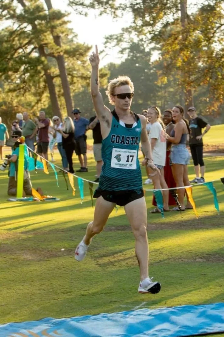 A male athlete wearing sunglasses and a blue and black tank top with the number 17 runs on a grassy field during a race, with people watching and trees in the background.