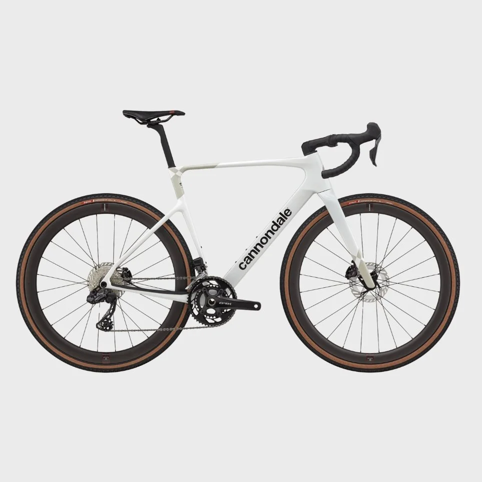 A white Cannondale gravel or road bicycle with black and tan tires, drop handlebars, and a black saddle, set against a plain light gray background.