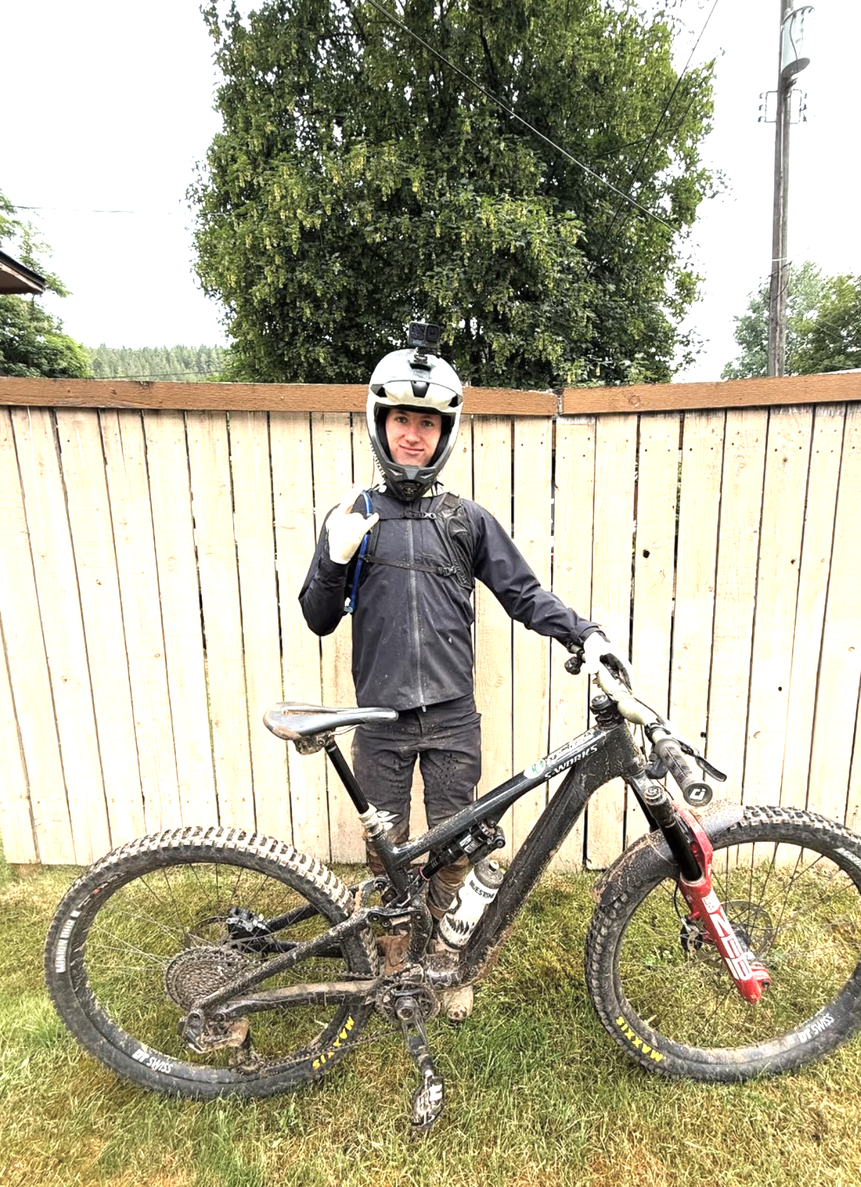 A person in mountain biking gear, including a helmet with a camera attached, standing with a muddy mountain bike in front of a wooden fence outdoors.