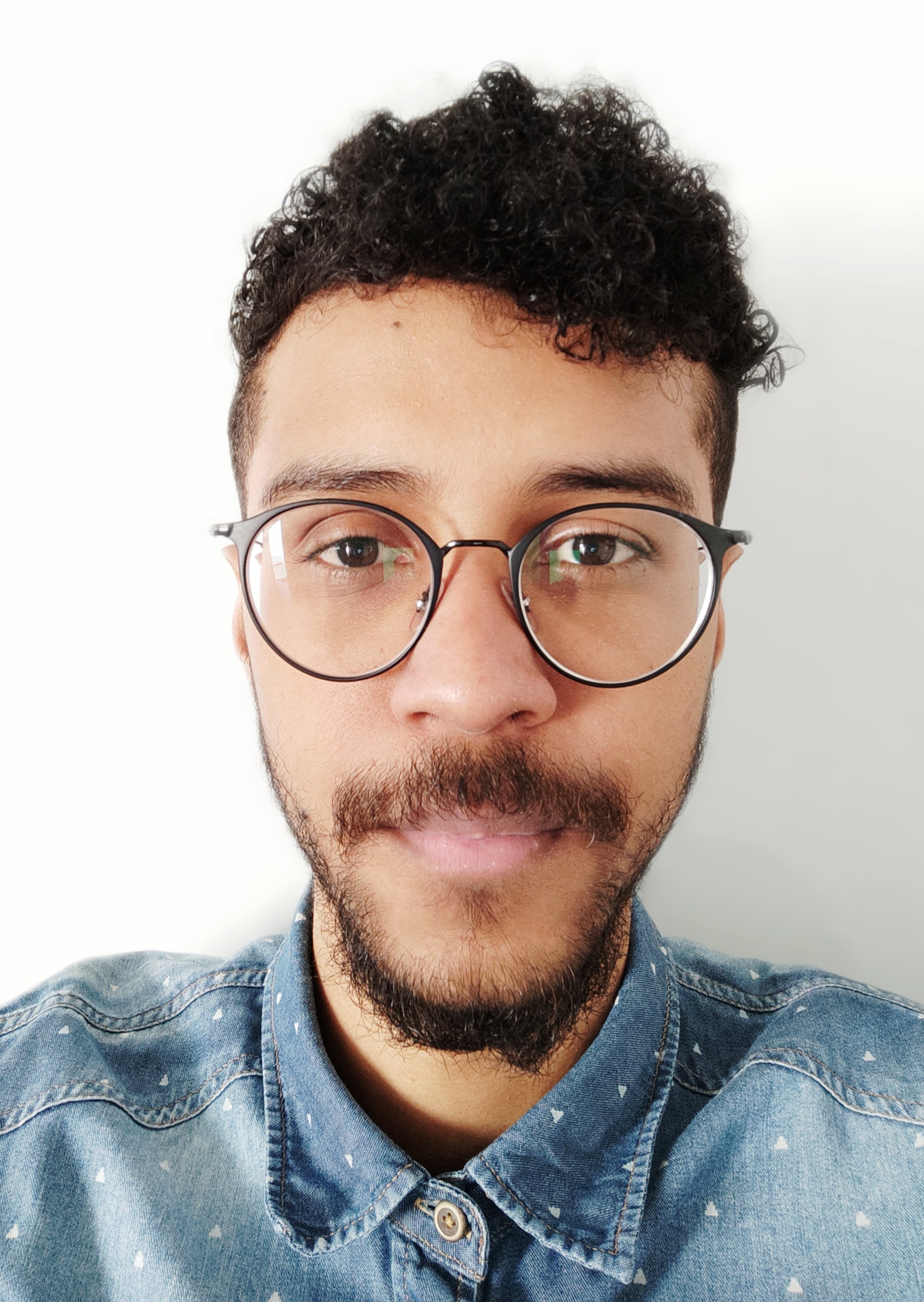 Close-up of a man with glasses, dark curly hair, and a beard, wearing a blue denim shirt.