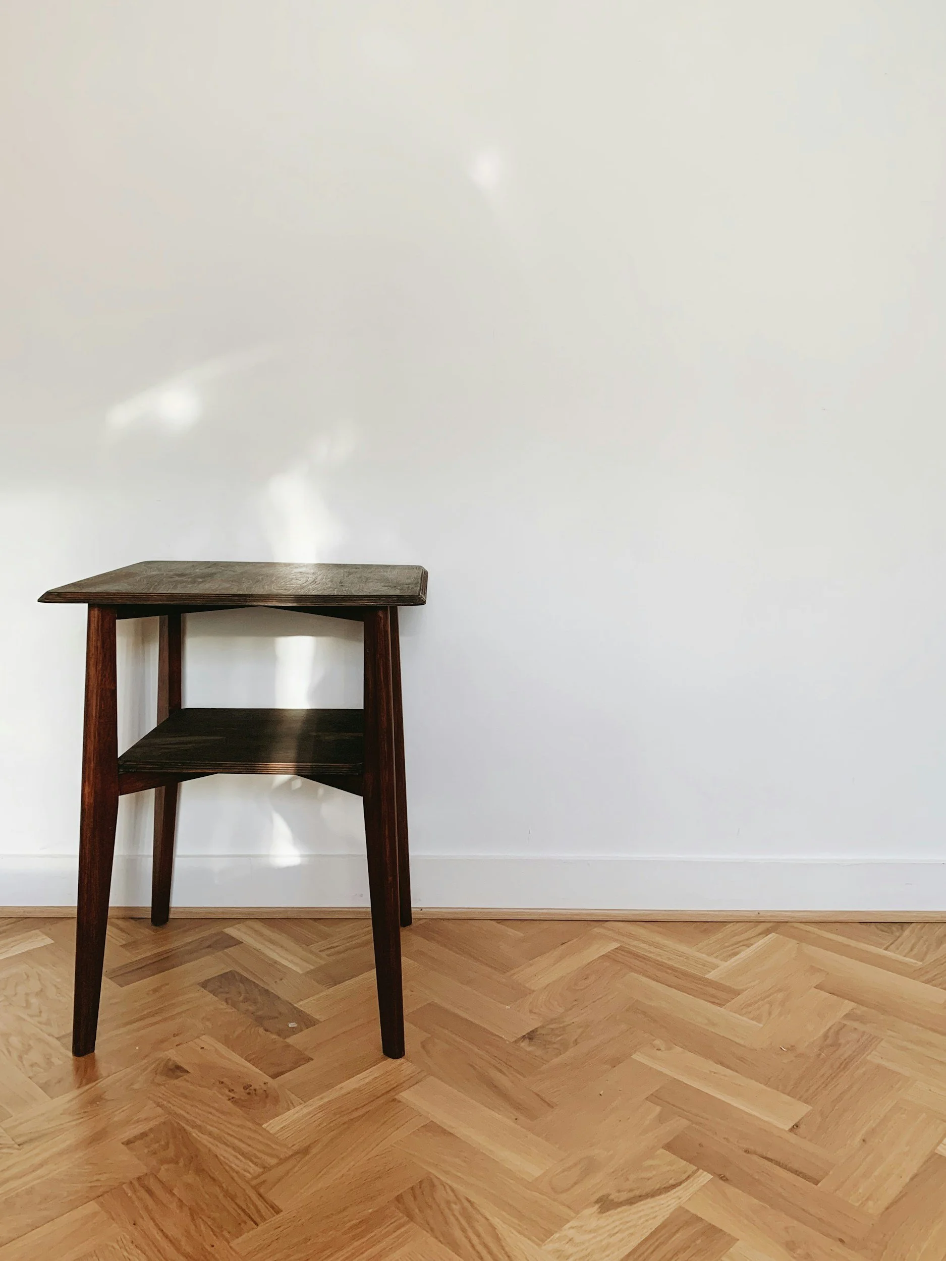 A dark wooden side table with a lower shelf against a plain white wall, sitting on a wooden parquet floor.