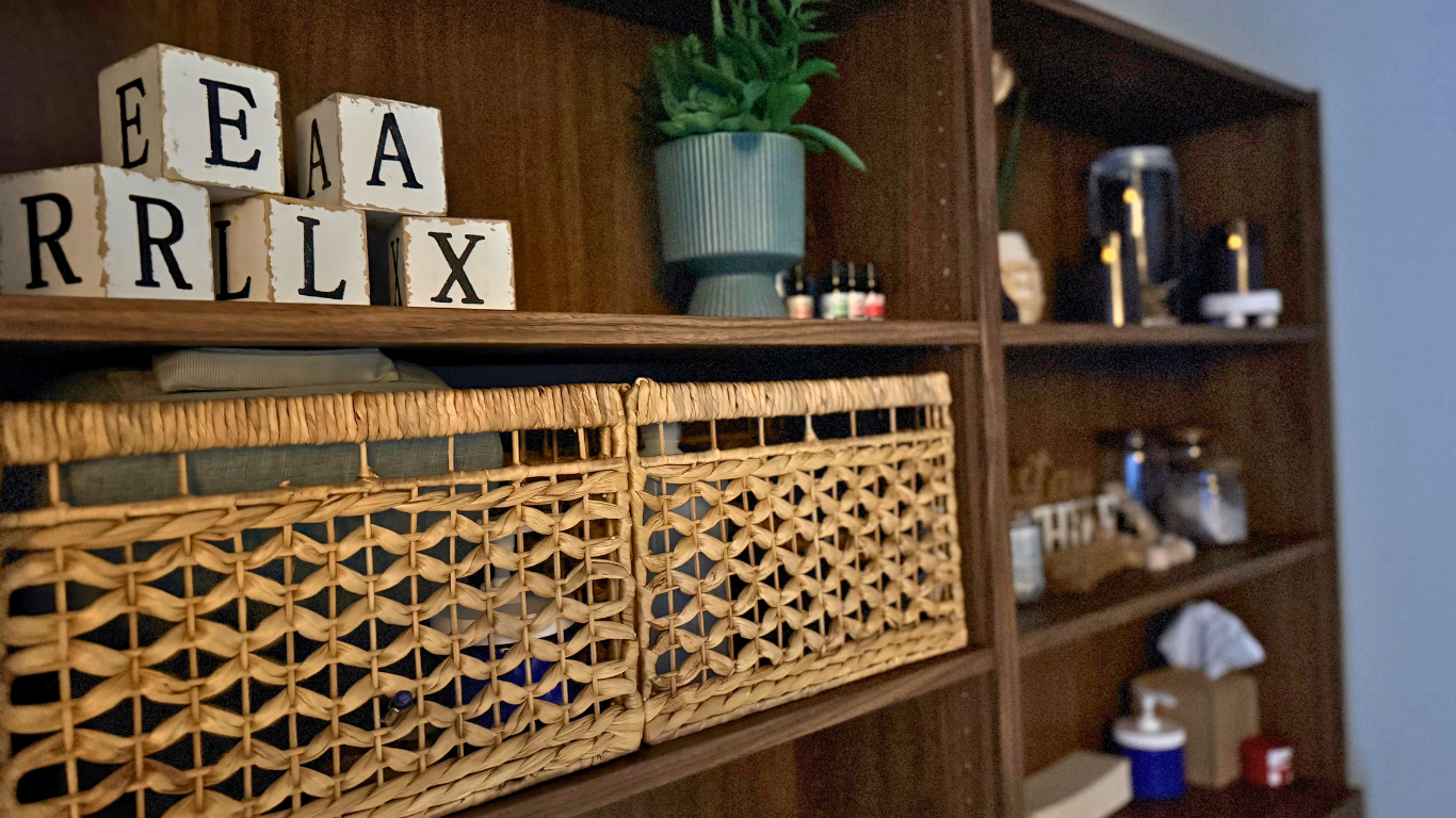 Decorative wooden bookshelf with variety of objects, including a green plant in a striped pot, herbal oils, and black bottles; woven baskets with fabric inside; wooden alphabet blocks spelling 'EARRX', 'A', 'L', 'R', 'X'; some shelves contain jars, a tissue box, and small miscellaneous items.