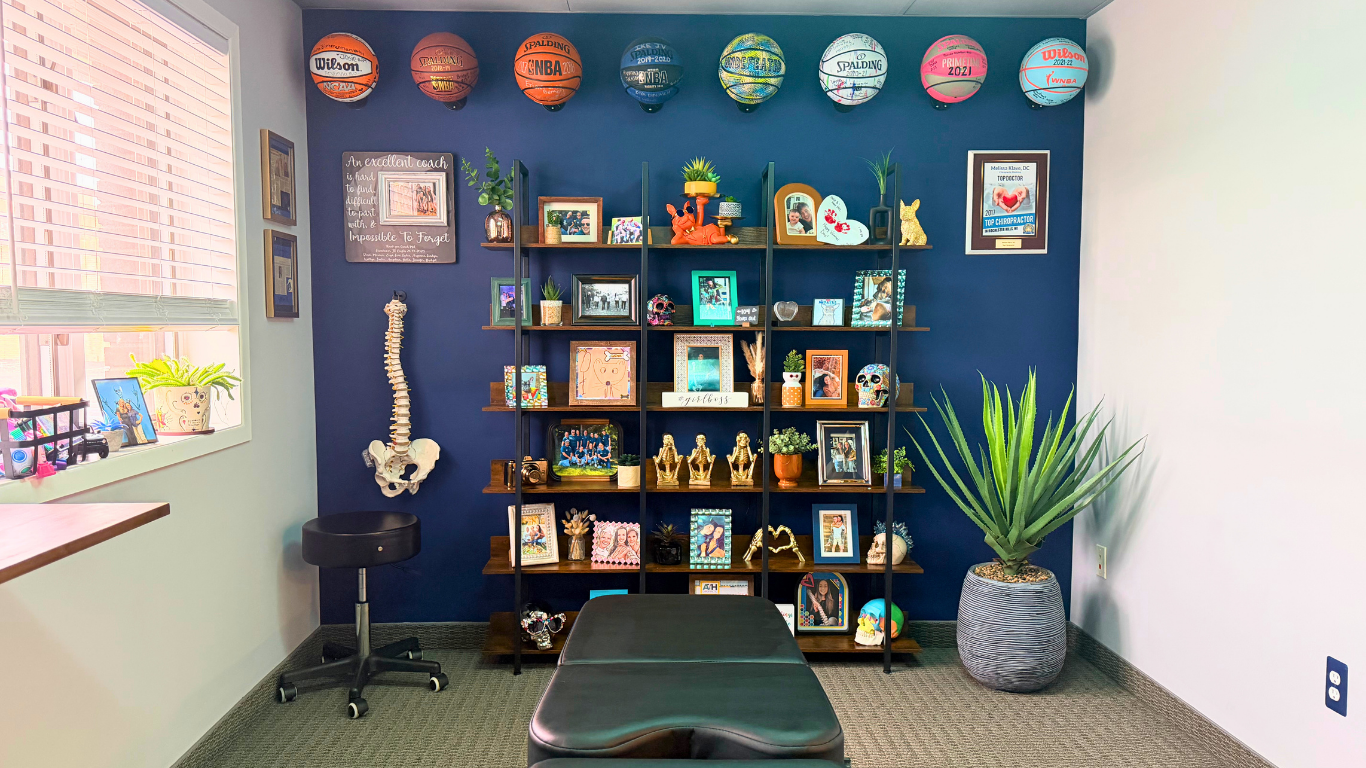 A room with a dark blue accent wall decorated with basketballs, framed pictures, sculptures, and plants. There is a large potted plant to the right and a window with blinds on the left side. A black stool and a black massage table are in the room.