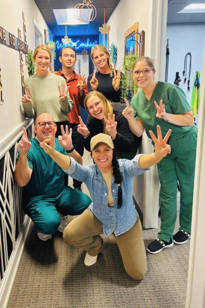 Group of seven people, including healthcare workers in scrubs, posing in a hallway with peace signs, smiling in a cheerful and celebratory mood.