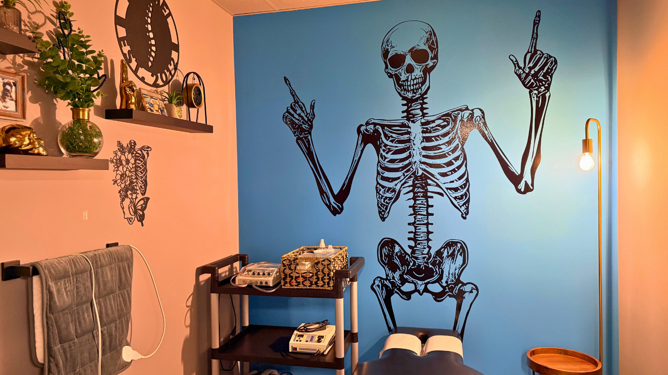 A blue wall with a large black skeleton mural holding up both middle fingers. In front, there's a massage table with towels, a small black cart with a phone and other items, and a wooden side table with a round tray. To the right, a floor lamp with a glowing bulb is visible, and to the left, shelves with plants, a clock, and decorative items are mounted on a pink wall.