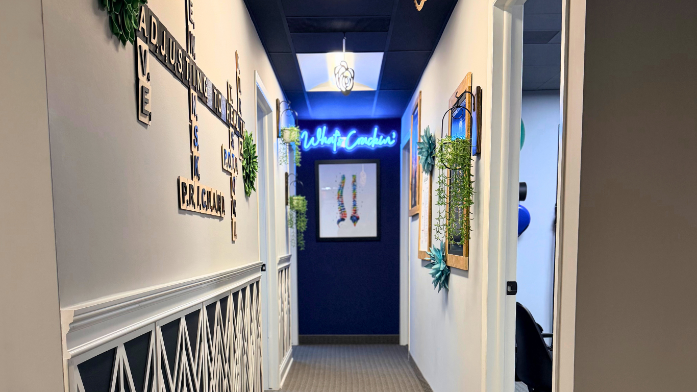 A hallway with white walls and a dark blue ceiling. On the left wall, there is black lettering art with words like 'Adjusting to Health', 'Inspire', 'Speak', and 'Breathe'. On the back wall, there is a framed picture and a blue neon sign that reads 'What's Cookin?'. The right wall has framed artwork and hanging plants, with a partially open door revealing a room with blue decor.