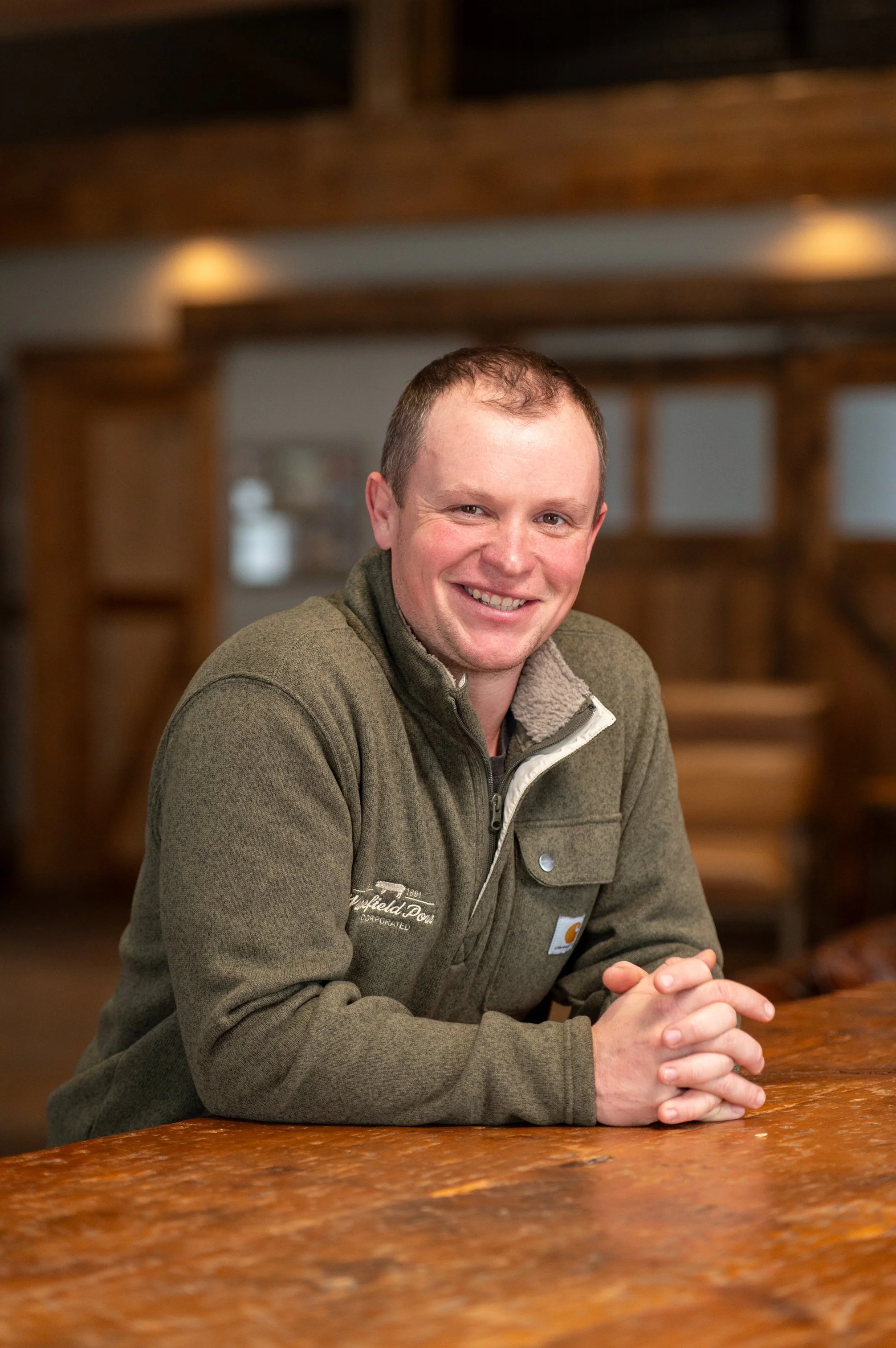 A smiling man with short brown hair and a green jacket sitting at a rustic wooden table in a warmly lit indoor space.