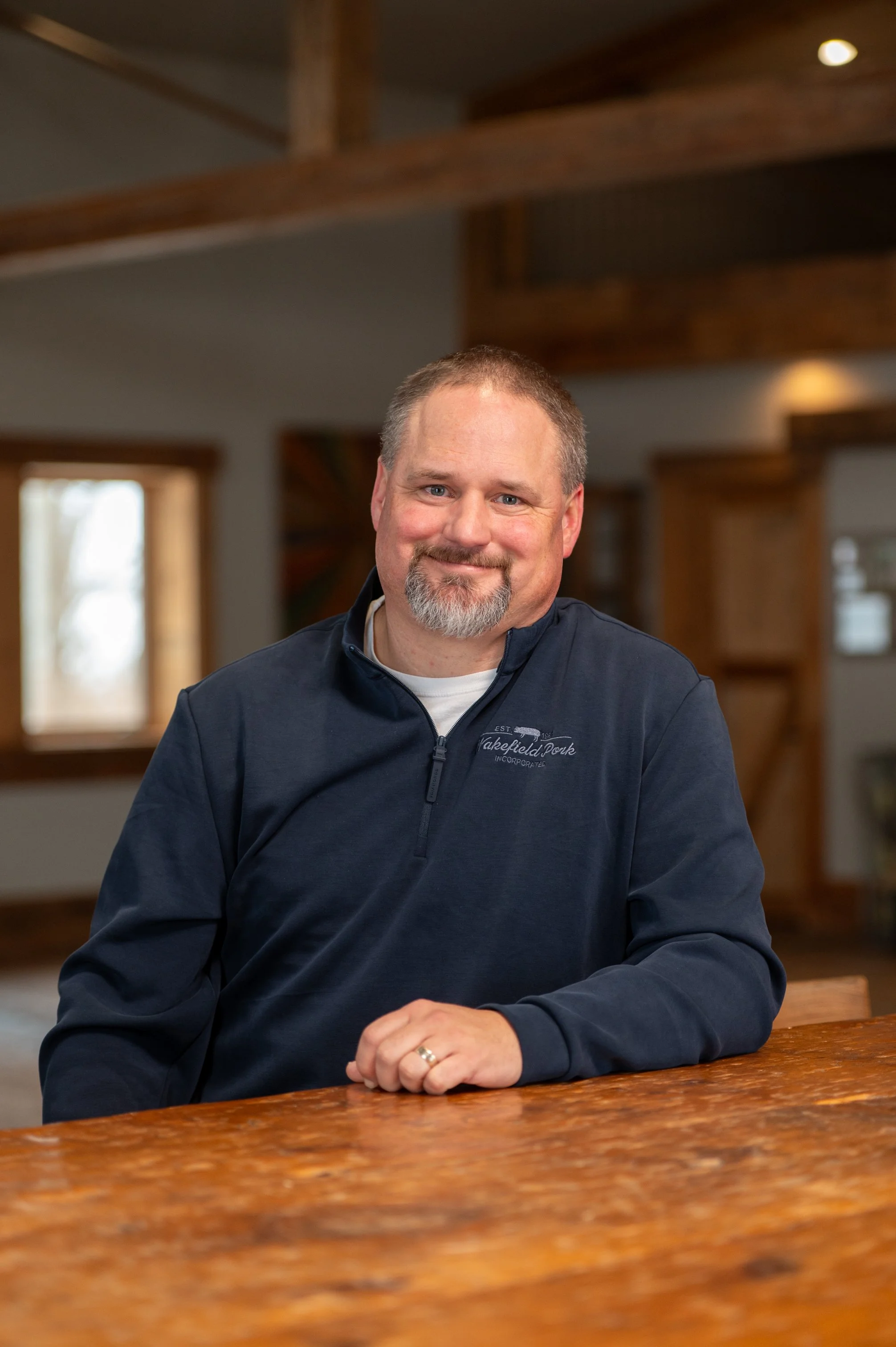 A middle-aged man with short hair, a beard, and a mustache, smiling and sitting at a wooden table in a warmly lit room with wooden beams and large windows in the background.