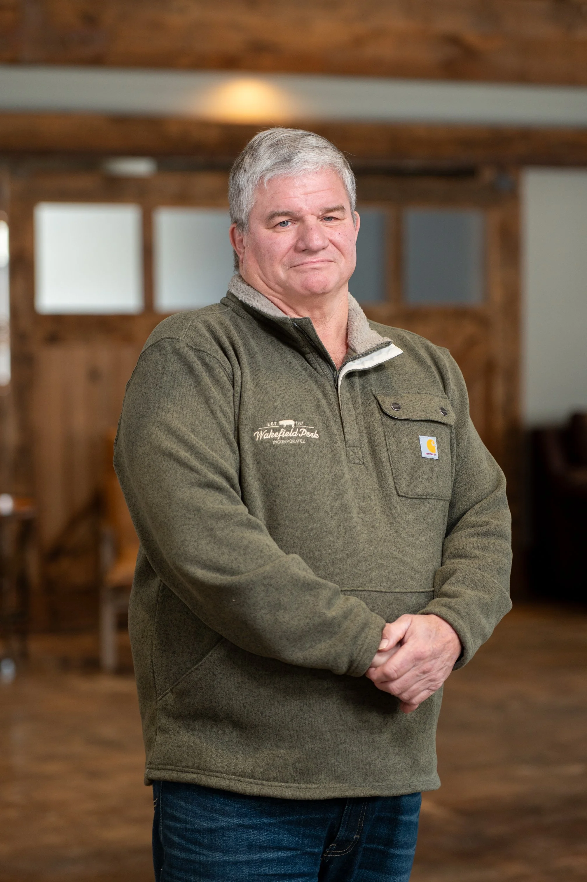 A middle-aged man with gray hair standing indoors in front of a wooden background. He is wearing a green fleece quarter-zip jacket with a Carhartt logo and a patch that says Nakefield Darek Inc. with his hands clasped in front of him.