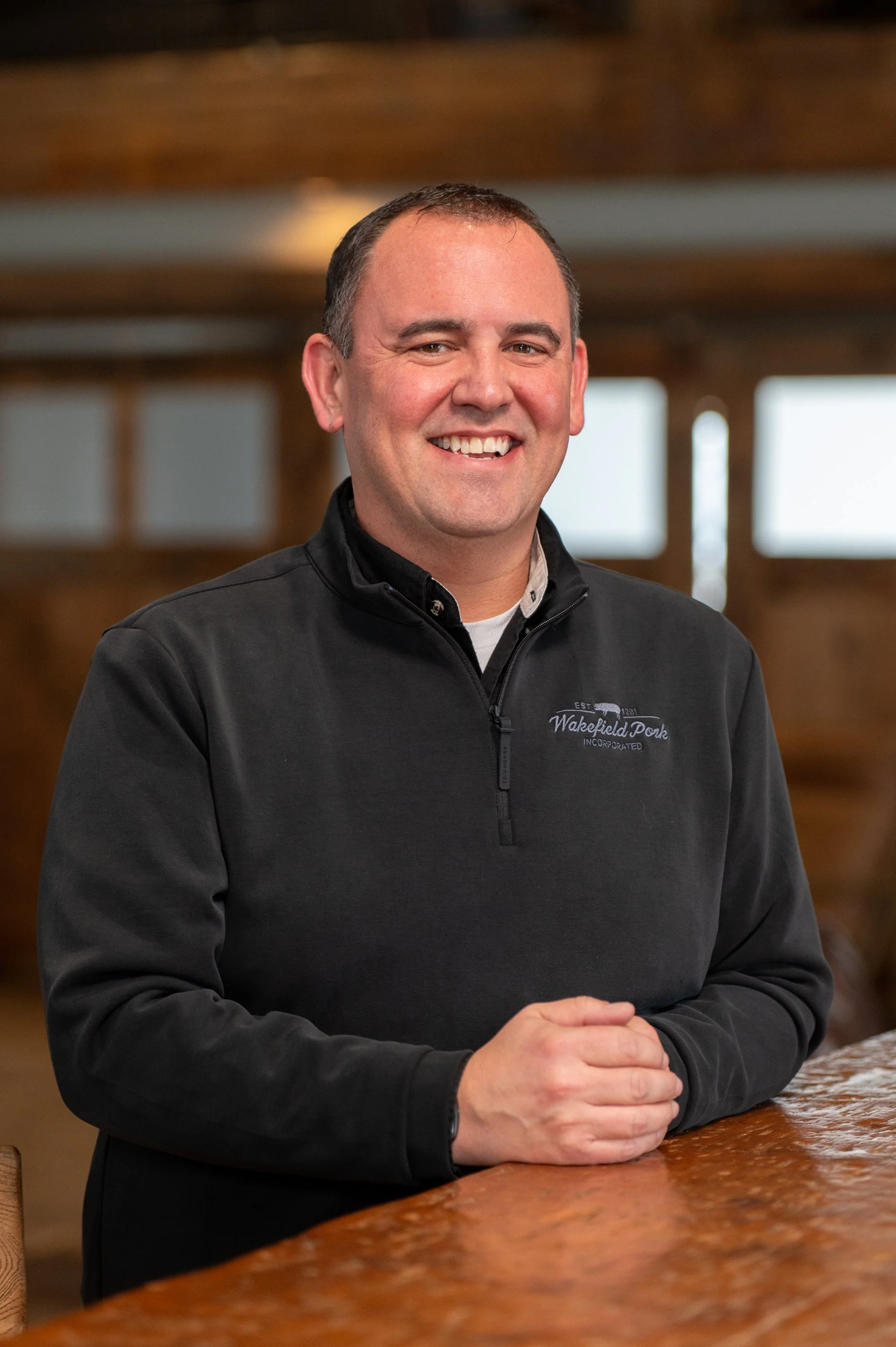A smiling man with short dark hair and light skin, wearing a black quarter-zip jacket with an embroidered logo that says 'Wakefield Dork Incorporated.' He is standing indoors in front of a wooden background, leaning on a wooden counter with his hands clasped together.