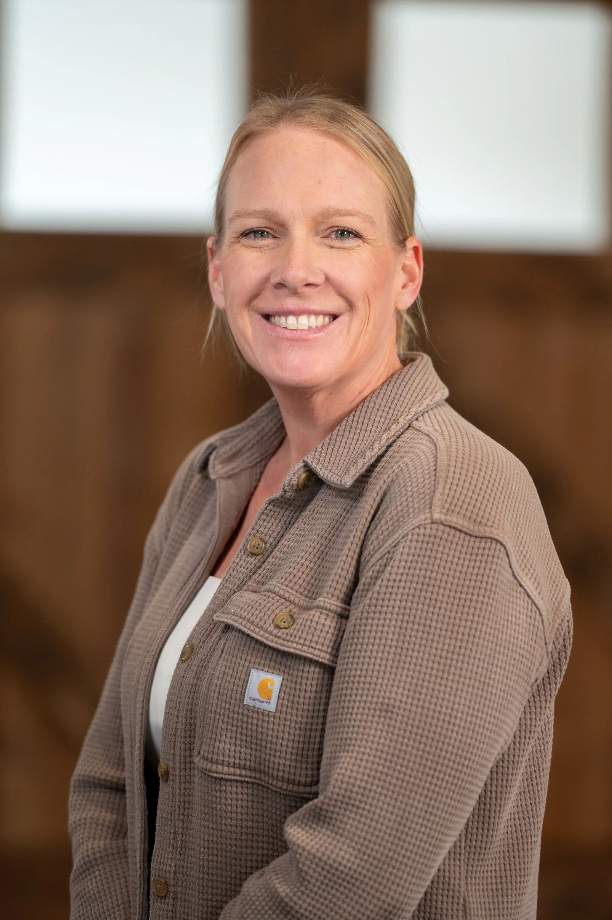 A smiling woman with blonde hair tied back, wearing a beige Carhartt jacket over a white top, standing indoors with a blurred background.