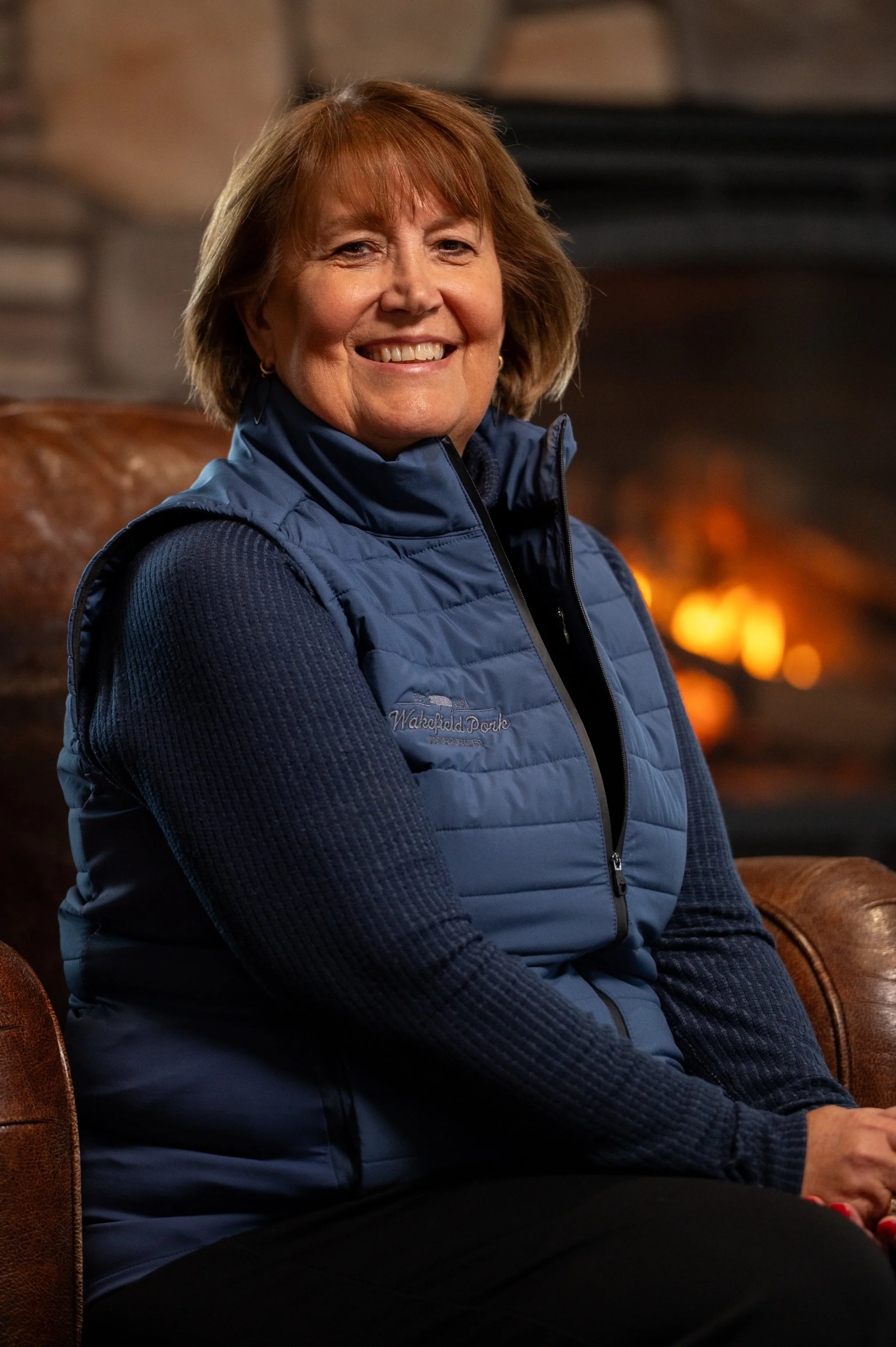 A smiling woman with short reddish-brown hair wearing a blue quilted vest and a navy blue sweater, sitting on a leather chair near a fireplace.