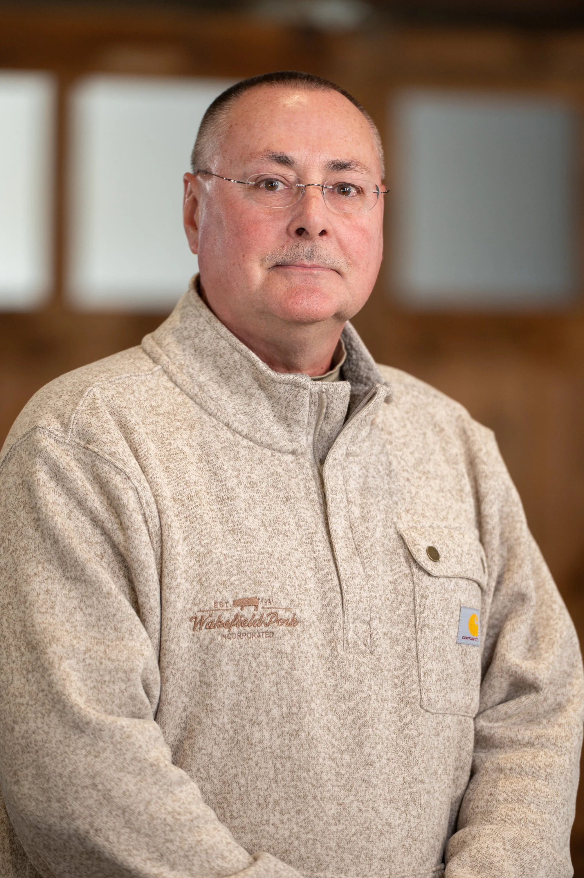 A middle-aged man with glasses and a shaved head, wearing a beige Carhartt fleece pullover with a small logo patch on the chest and a pocket, standing indoors with a blurred wooden background.