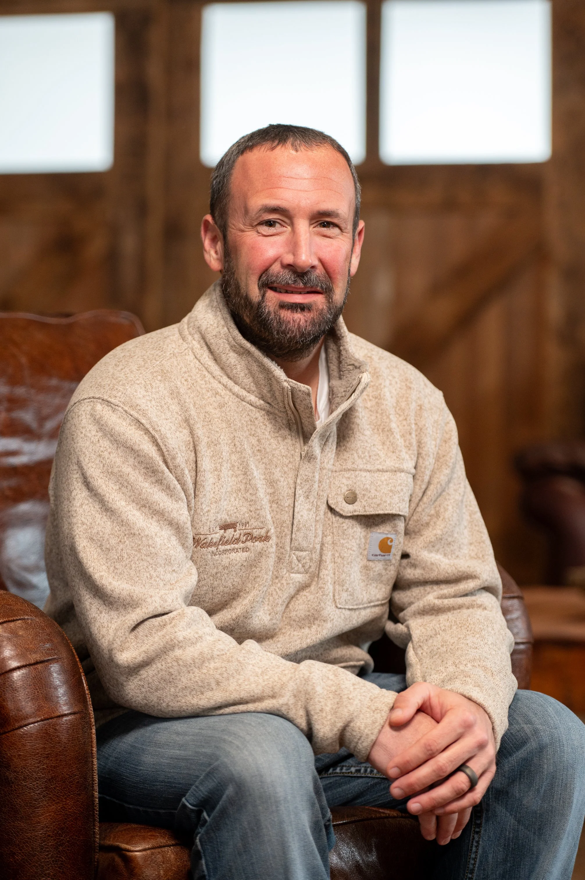 A man with a beard and short hair, wearing a beige Carhartt fleece jacket, sitting on a brown leather chair in a rustic wooden building with large windows.