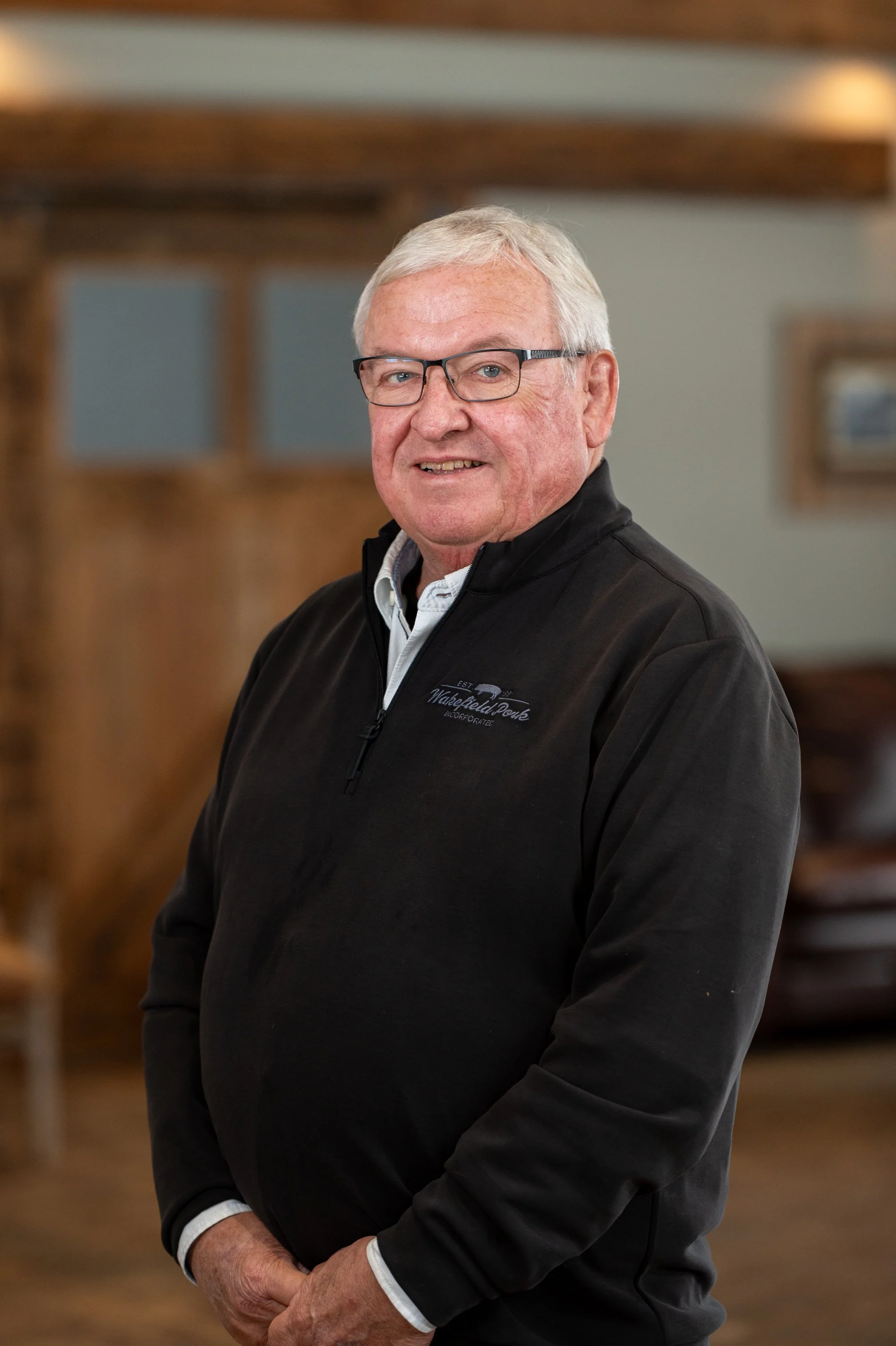 A senior man with glasses and gray hair, wearing a black zip-up jacket over a collared shirt, standing indoors in front of a wooden staircase and smiling at the camera.