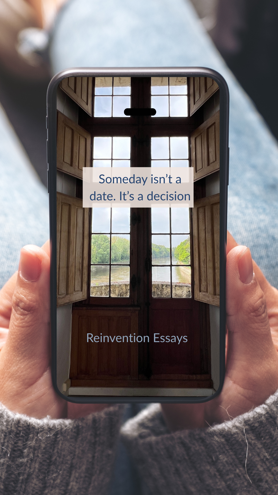 Person holding a phone displaying the cover of 'Reinvention Essays' with a window view in the background and an inspirational quote: 'Someday isn't a date. It's a decision.'