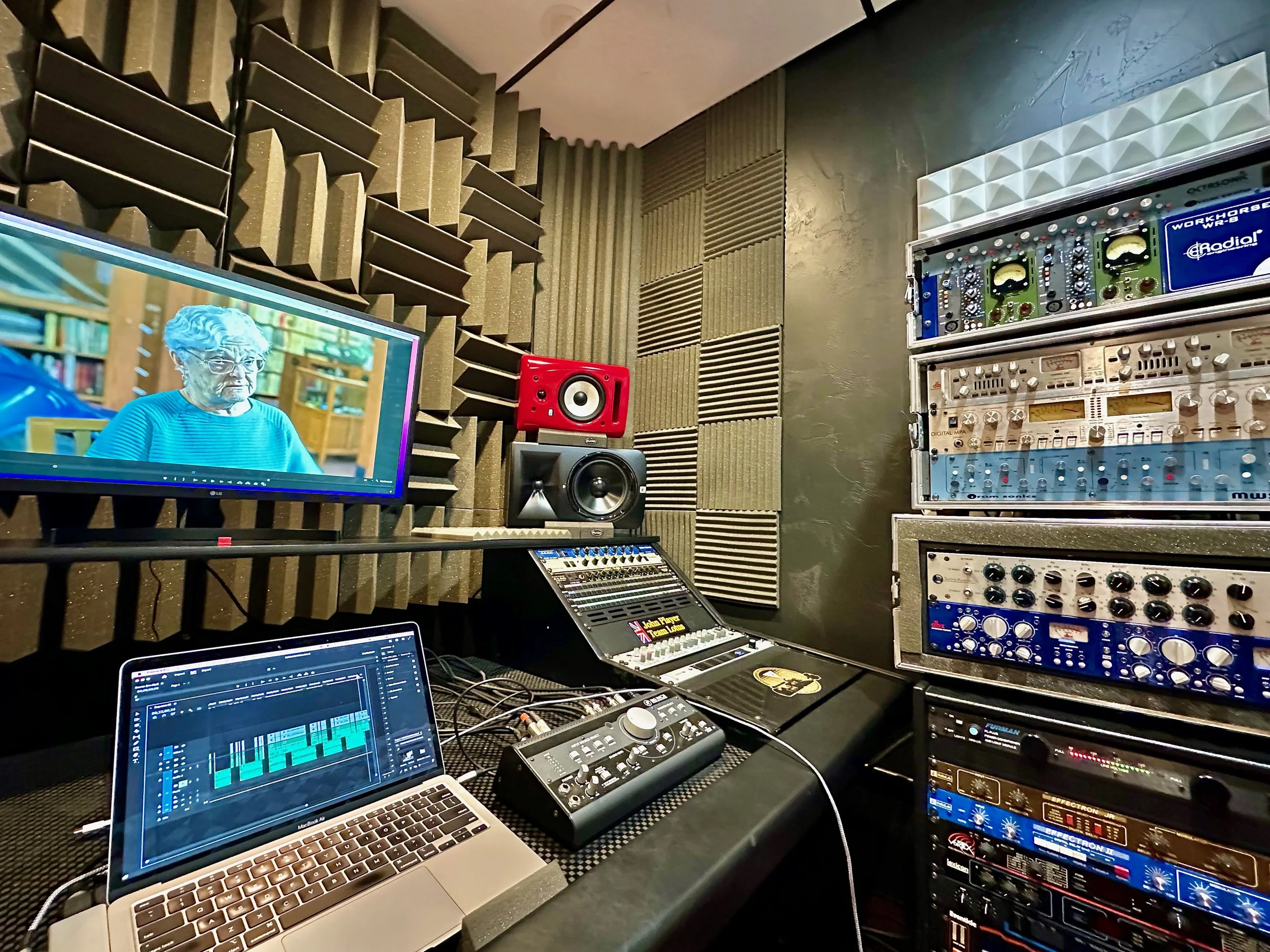 Audio recording studio with soundproof foam walls, a computer with music production software, studio monitors, audio mixers, and racks of audio equipment.