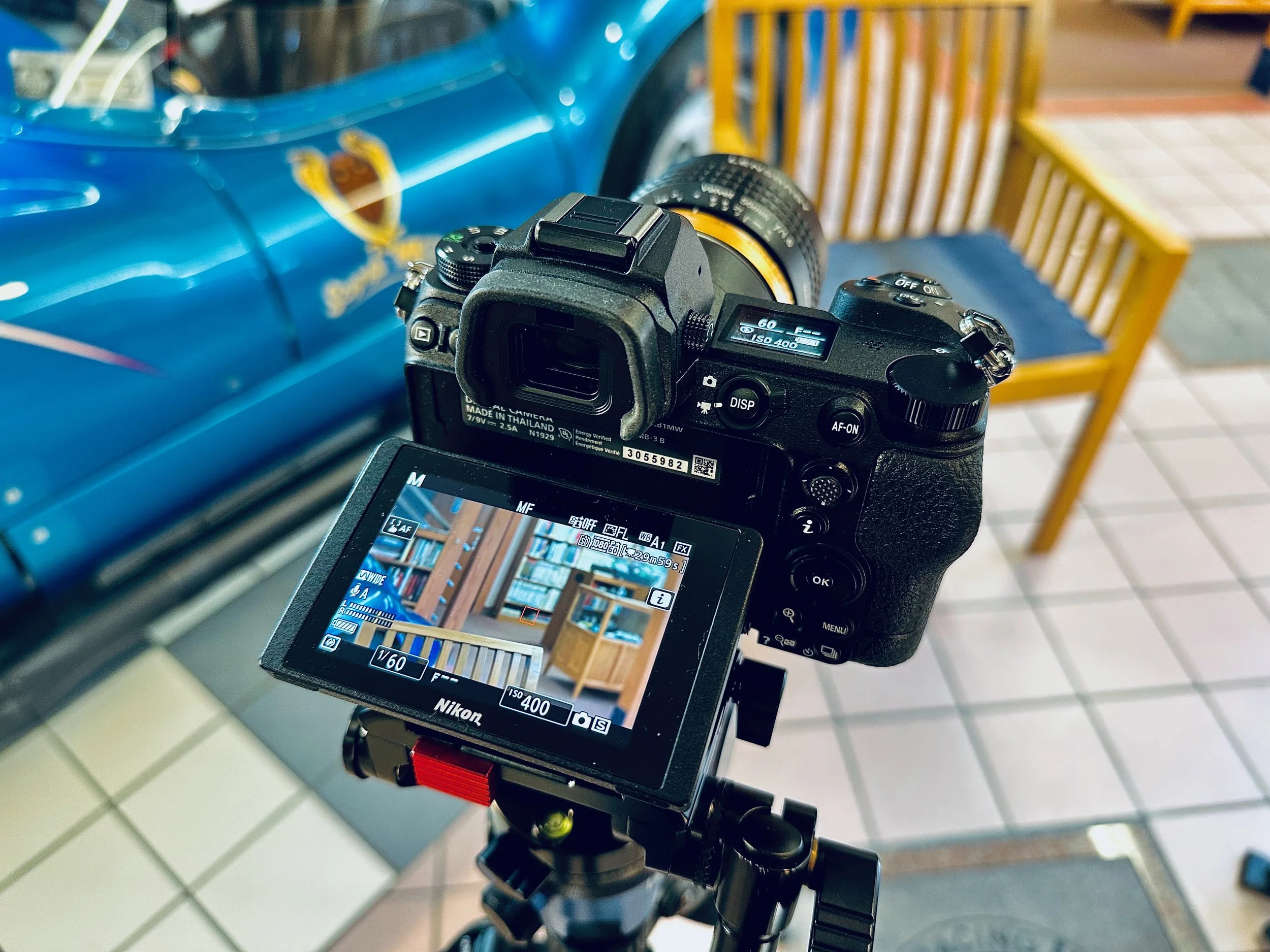A camera on a tripod capturing a photo of a vintage blue race car with a yellow crest, parked indoors with tiled flooring and a wooden bench in the background.