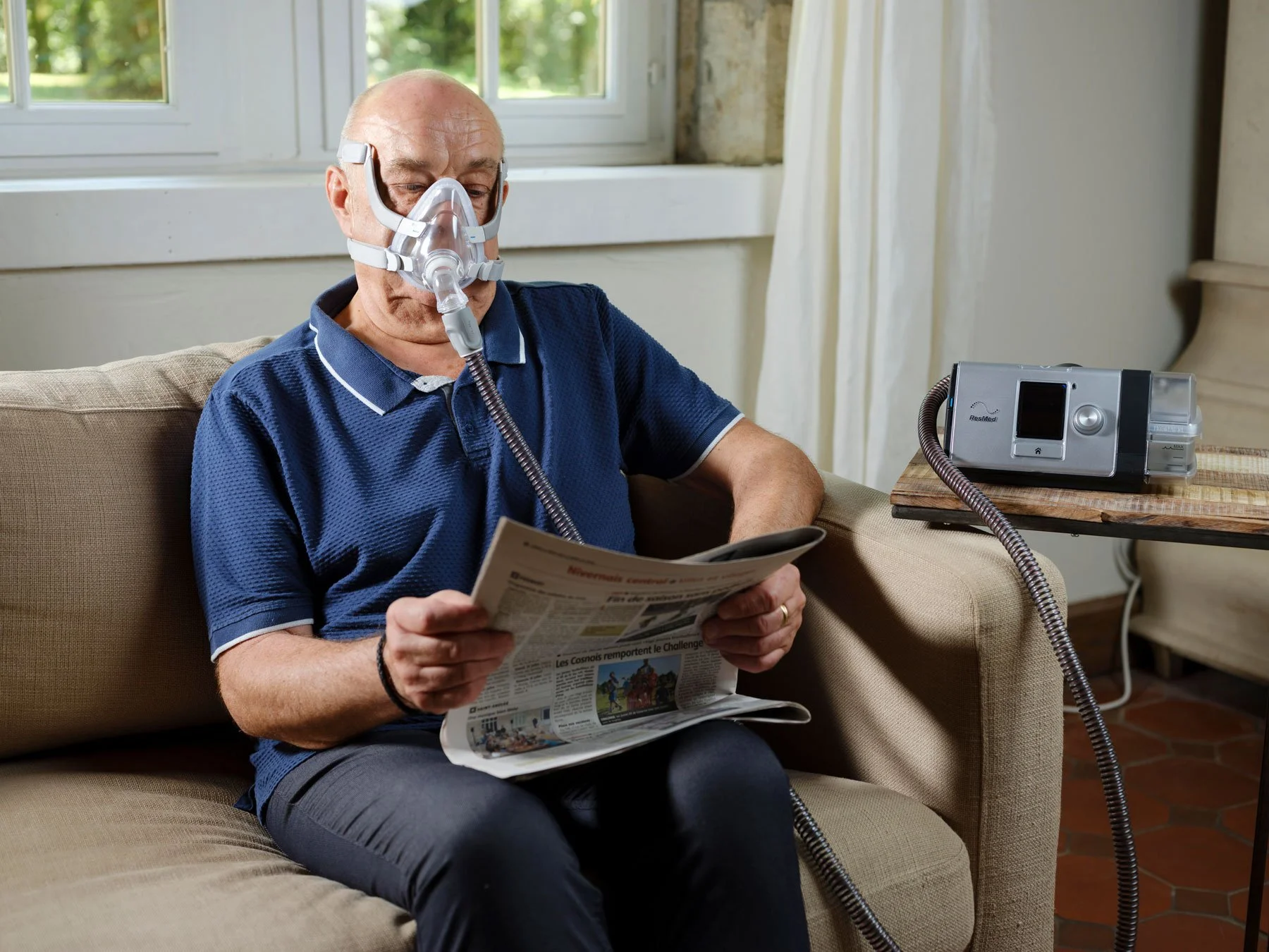 An elderly man sitting on a beige couch wearing a CPAP mask connected to a CPAP machine placed on a wooden table nearby. He is reading a newspaper in a well-lit room with a window behind him.