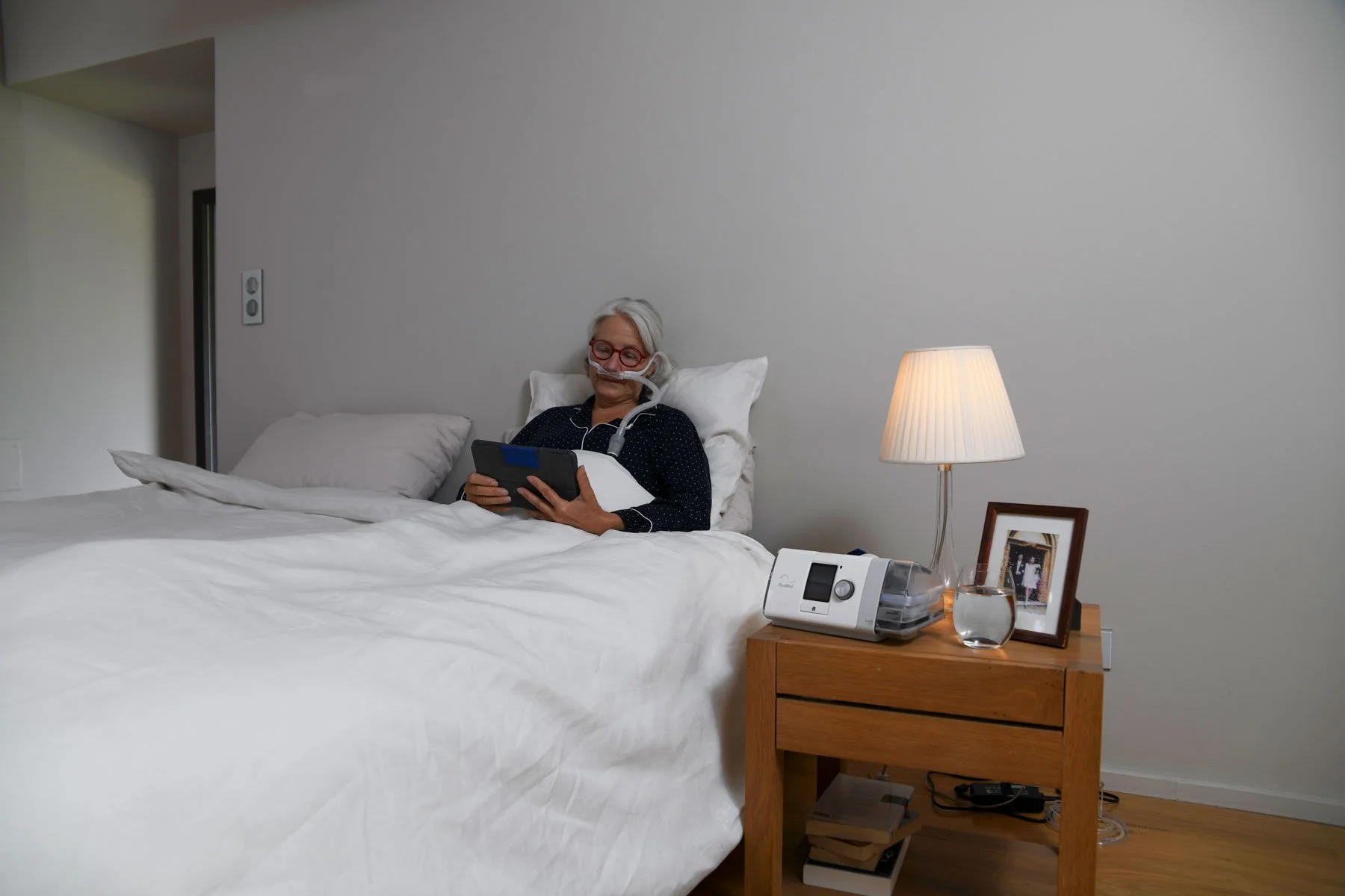 An elderly woman in bed with a nasal oxygen tube, holding a tablet, with an oxygen concentrator on the bedside table.