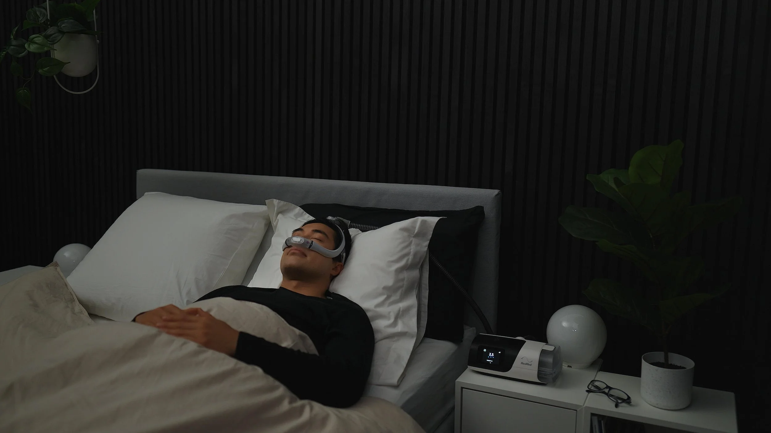 A man sleeping on a bed with a sleep apnea CPAP machine next to him, wearing a sleep apnea mask, in a dimly lit bedroom.