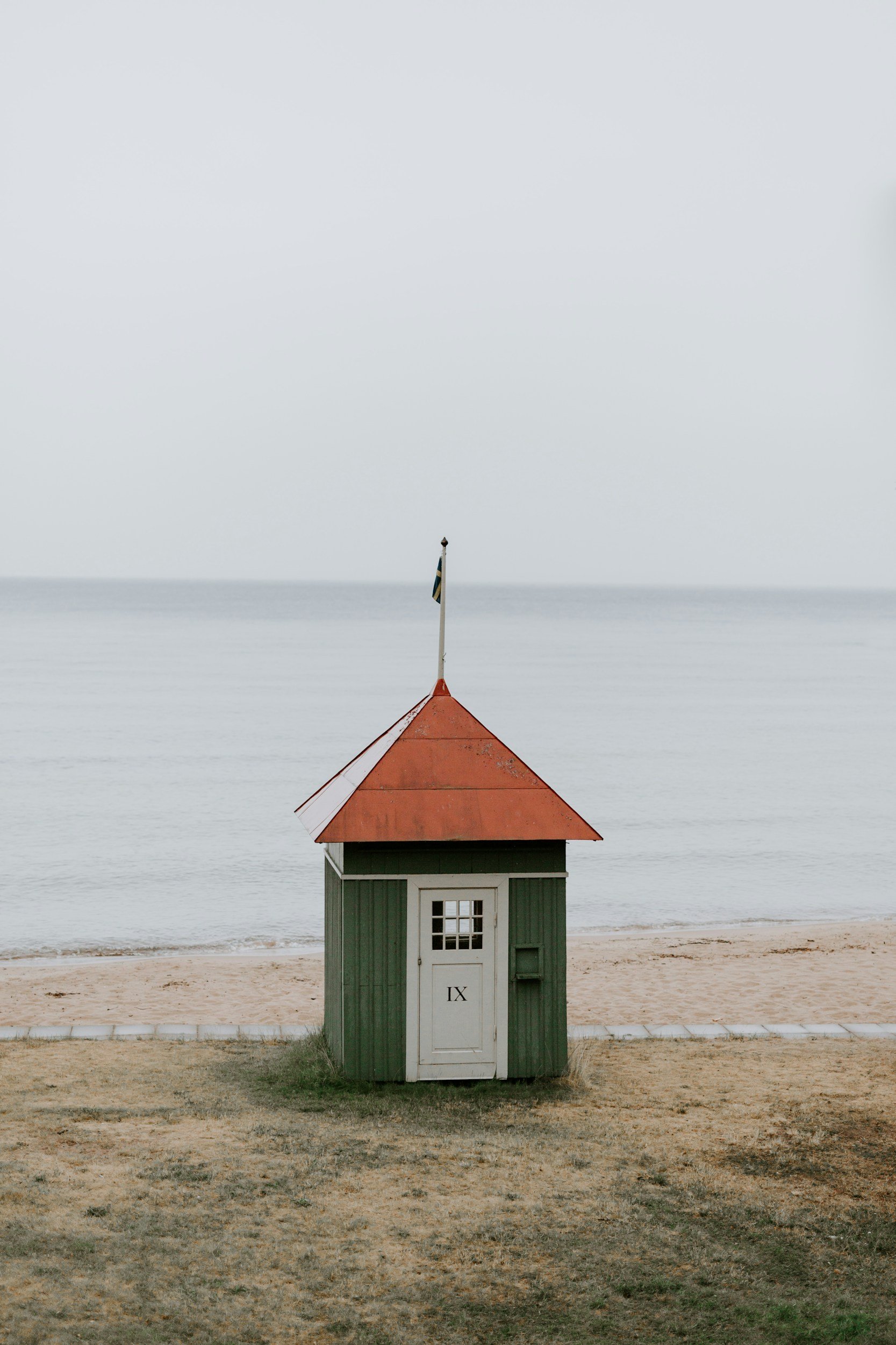 Small green wooden beach hut with a red shingle roof on sand, overlooking calm sea under overcast sky.