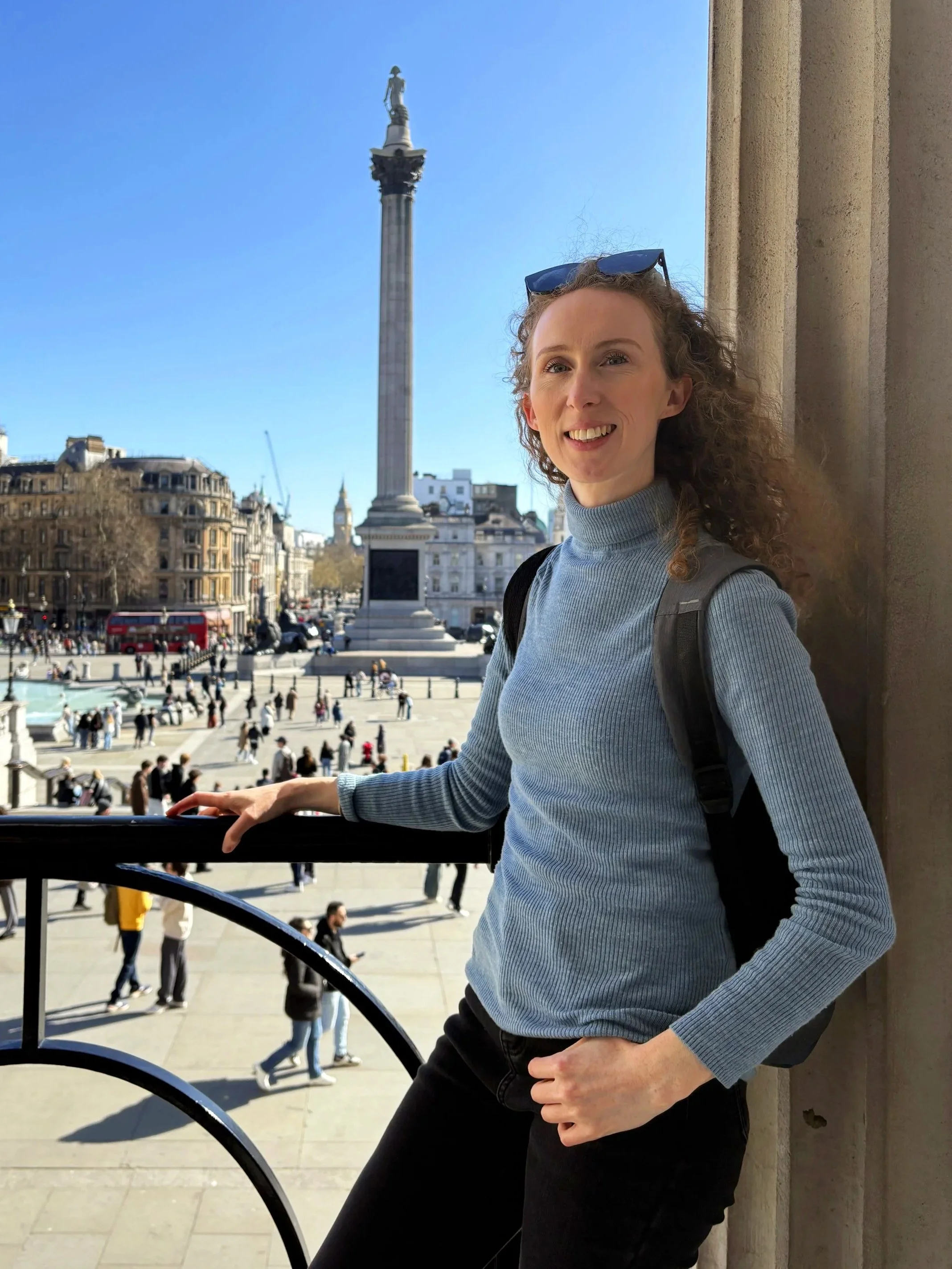 A woman with curly hair wearing a blue turtleneck sweater and sunglasses on her head, standing near a balcony railing in Trafalgar Square, London, with Nelson's Column and buildings in the background on a sunny day.