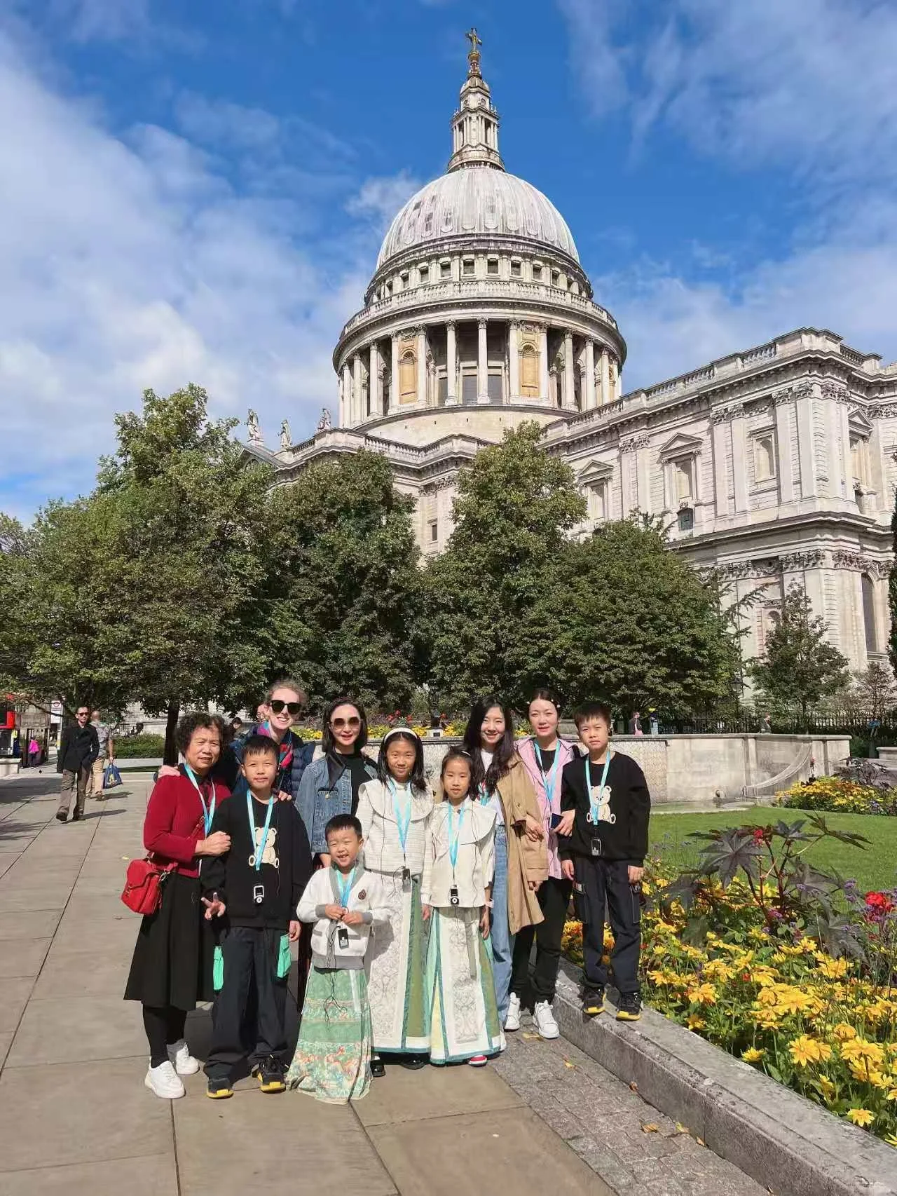 Group of children and adults standing in front of St. Paul's Cathedral in London, England, with a clear blue sky and colorful flowers in the foreground.