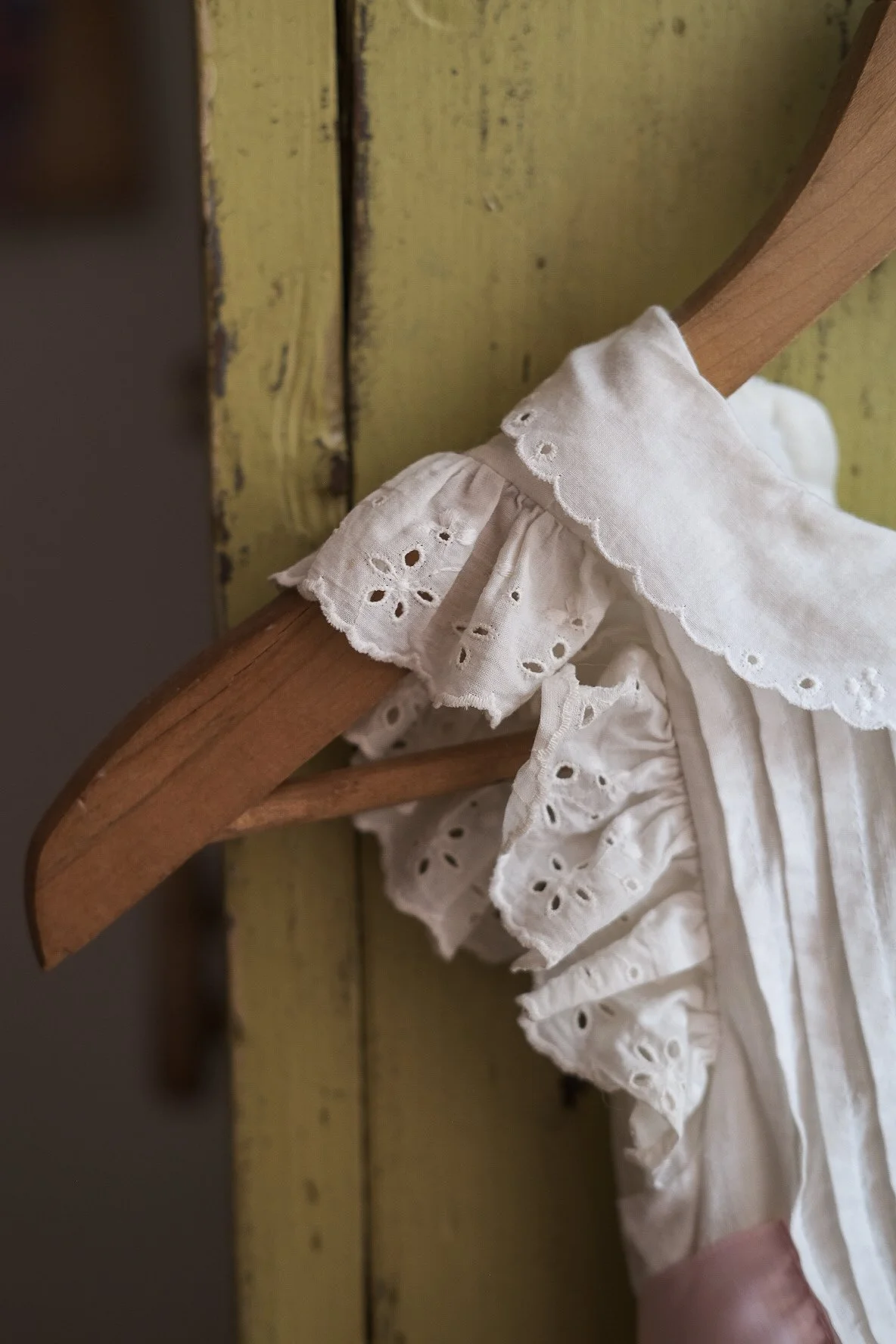Close-up of a wooden hanger with a white embroidered baby dress hanging on it, set against a rustic yellow wooden wall.