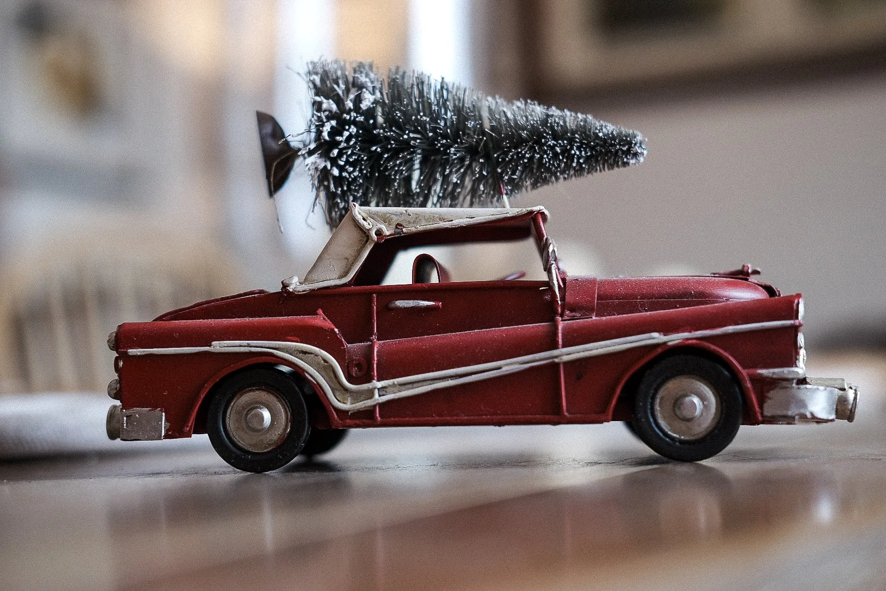 A vintage toy pickup truck in red with a white roof and trim, carrying a small artificial Christmas tree on top, placed on a flat surface.