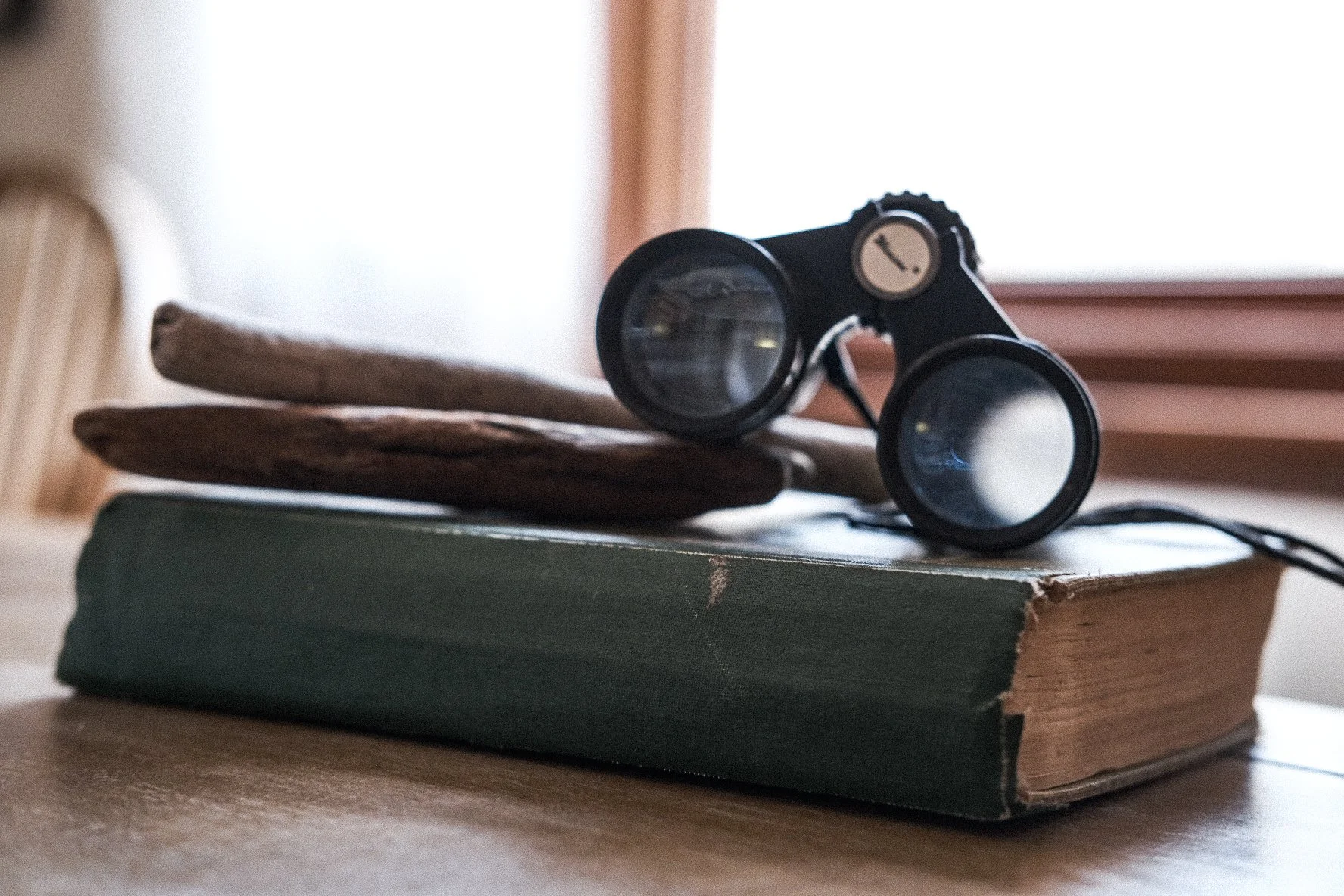 A pair of vintage binoculars resting on a stack of old books and wooden sticks, on a wooden surface near a window.