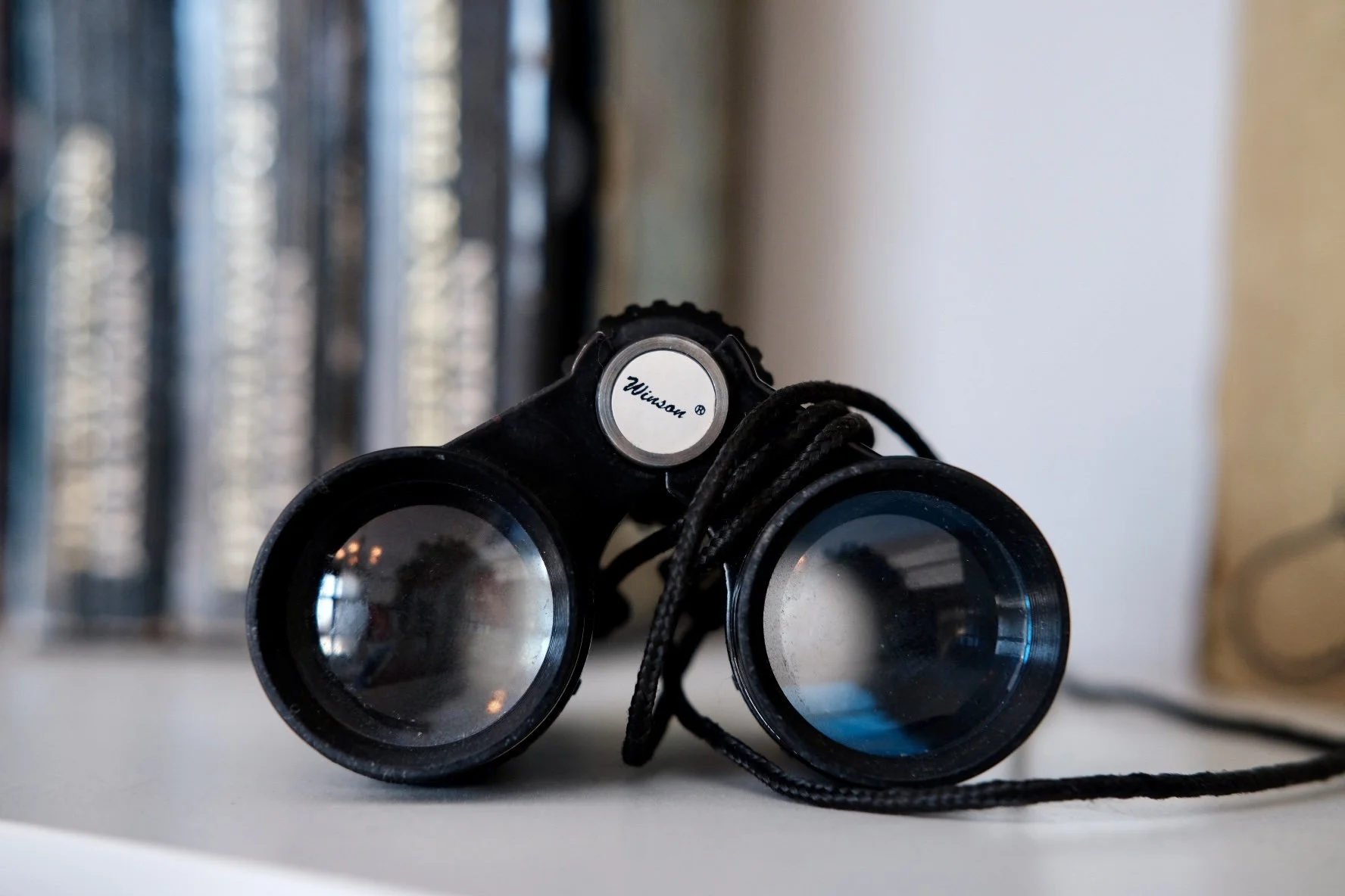 Pair of black binoculars with blue-tinted lenses resting on a white surface, with a blurred background of books or files on a shelf.