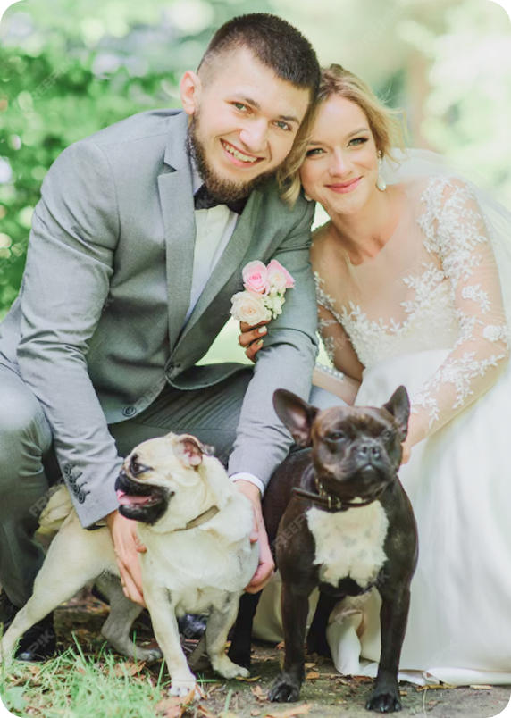 A smiling bride and groom in wedding attire with two dogs outdoors on a grassy area with trees in the background.