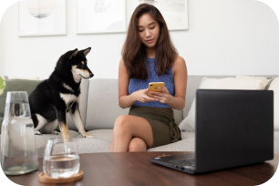 Woman sitting on a couch using a smartphone with a black and tan dog beside her and a laptop on a coffee table.