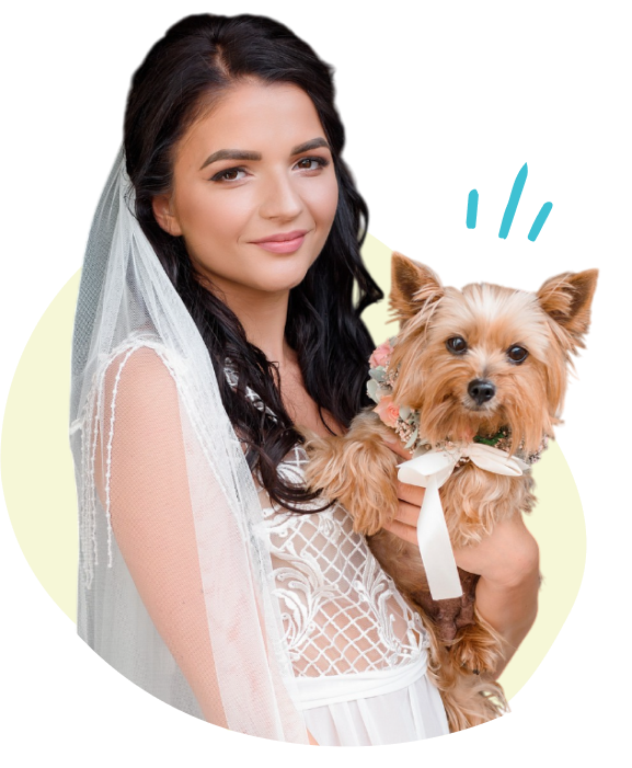 A woman dressed as a bride holding a small dog with a floral collar and a ribbon, posing against a plain background.