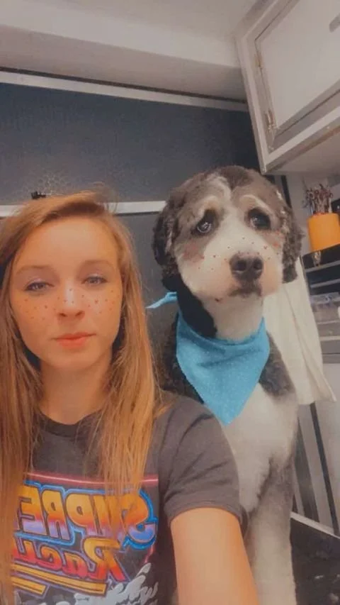 A young woman and a dog with facial filters, adding freckles to both of them, in an indoor setting. The dog is wearing a blue bandana.