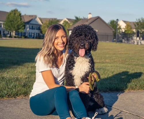 Woman sitting on grass with a large black and white dog holding a toy against a suburban backdrop.